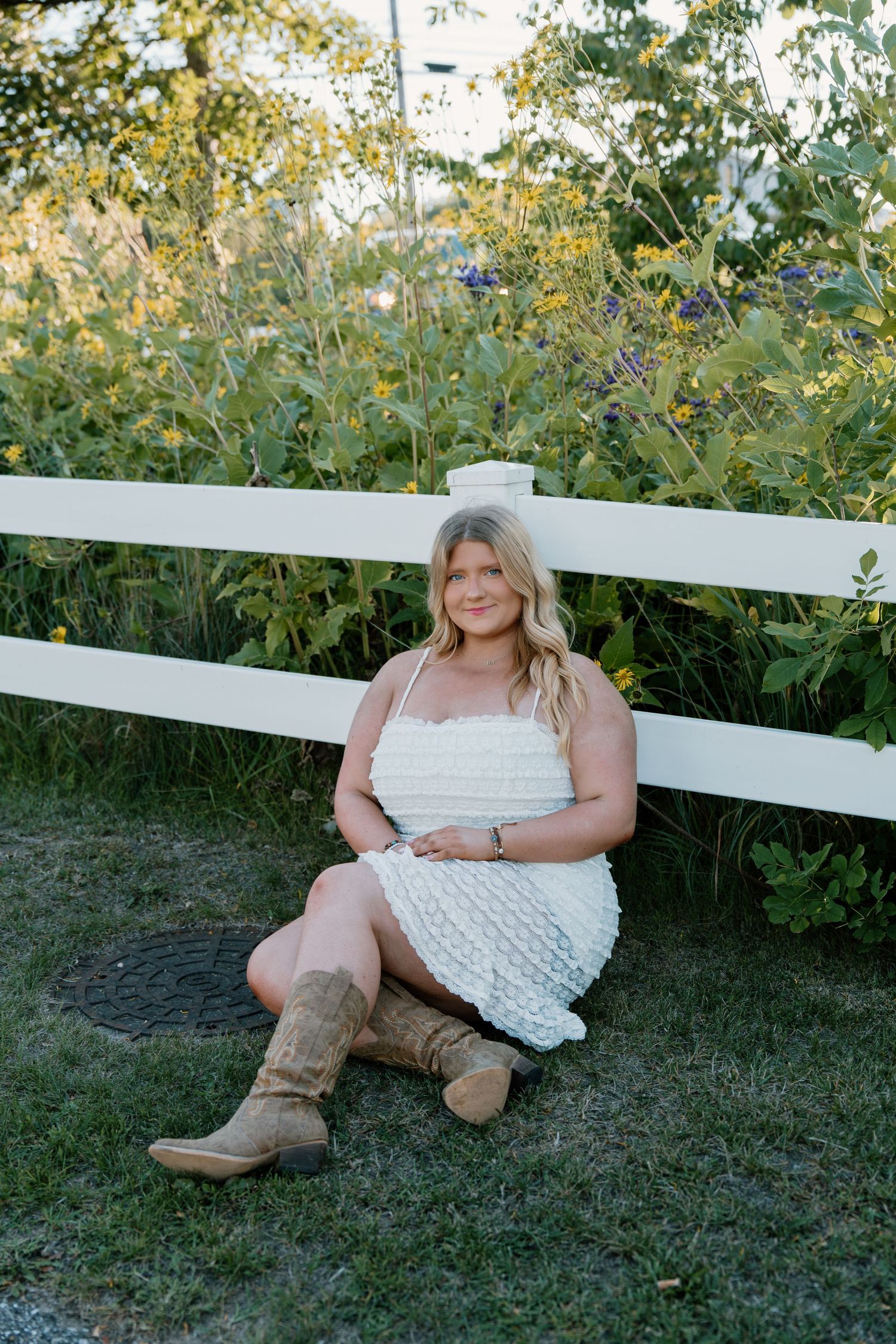 Young woman in white dress and cowboy boots sitting by white fence surrounded by wildflowers.
