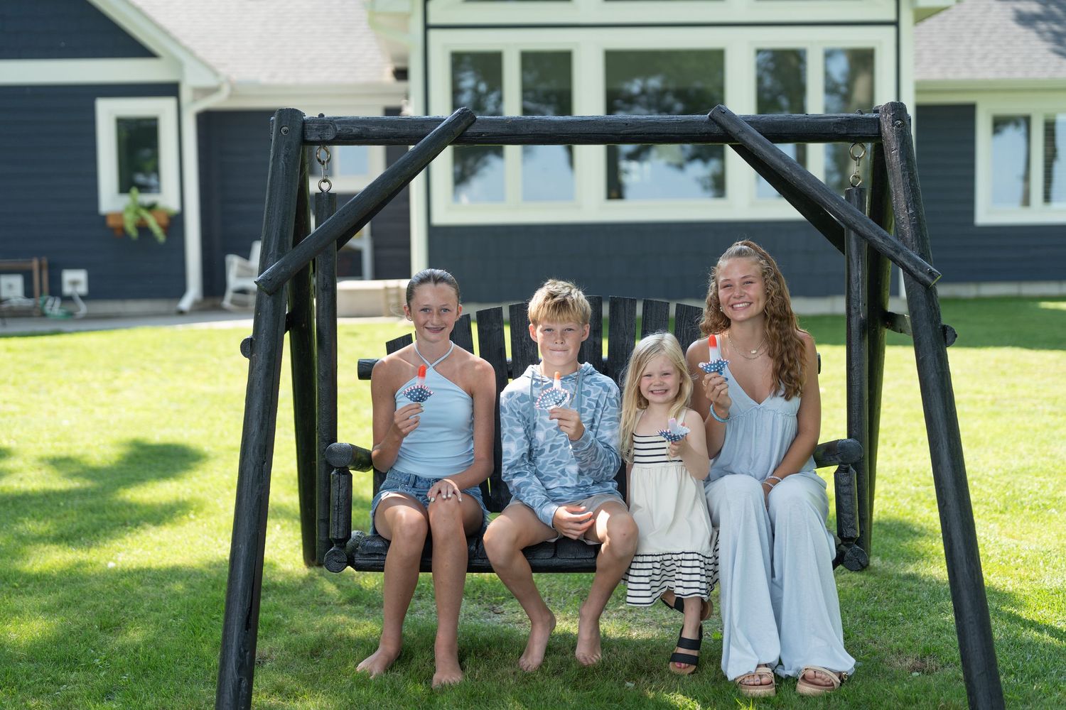 Four children smiling on a black wooden porch swing in a sunny backyard, holding popsicles on a summer day.
