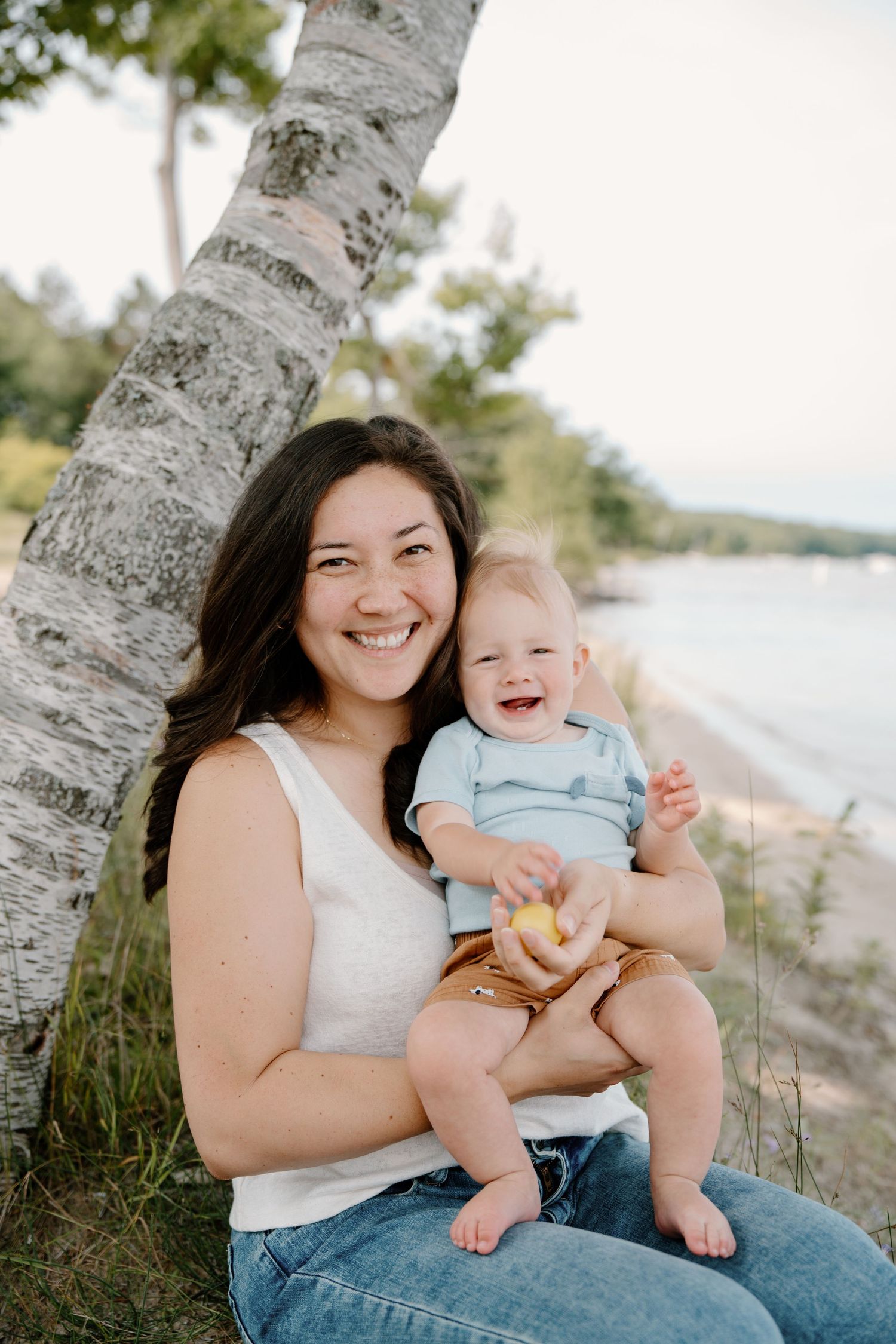 Smiling woman holding a happy baby boy near a birch tree on a scenic beach shoreline.