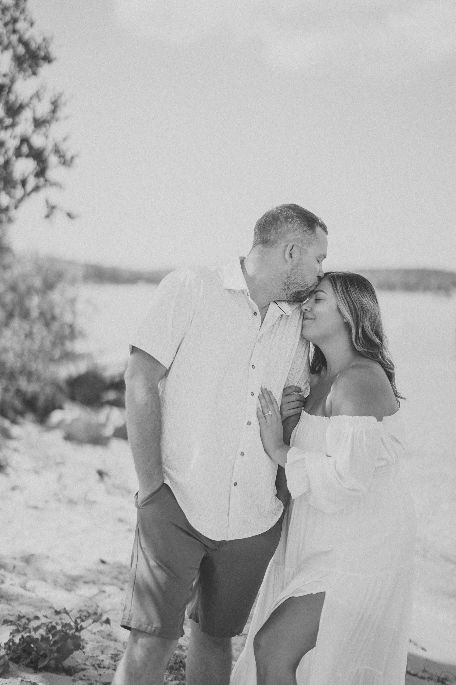 A romantic black and white photo of a couple sharing an intimate moment on a lakeside beach.