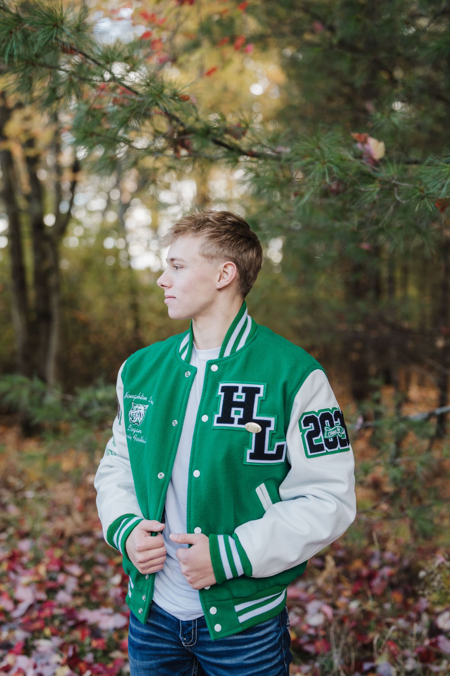 Person in green and white letterman jacket poses among autumn trees and fallen leaves.