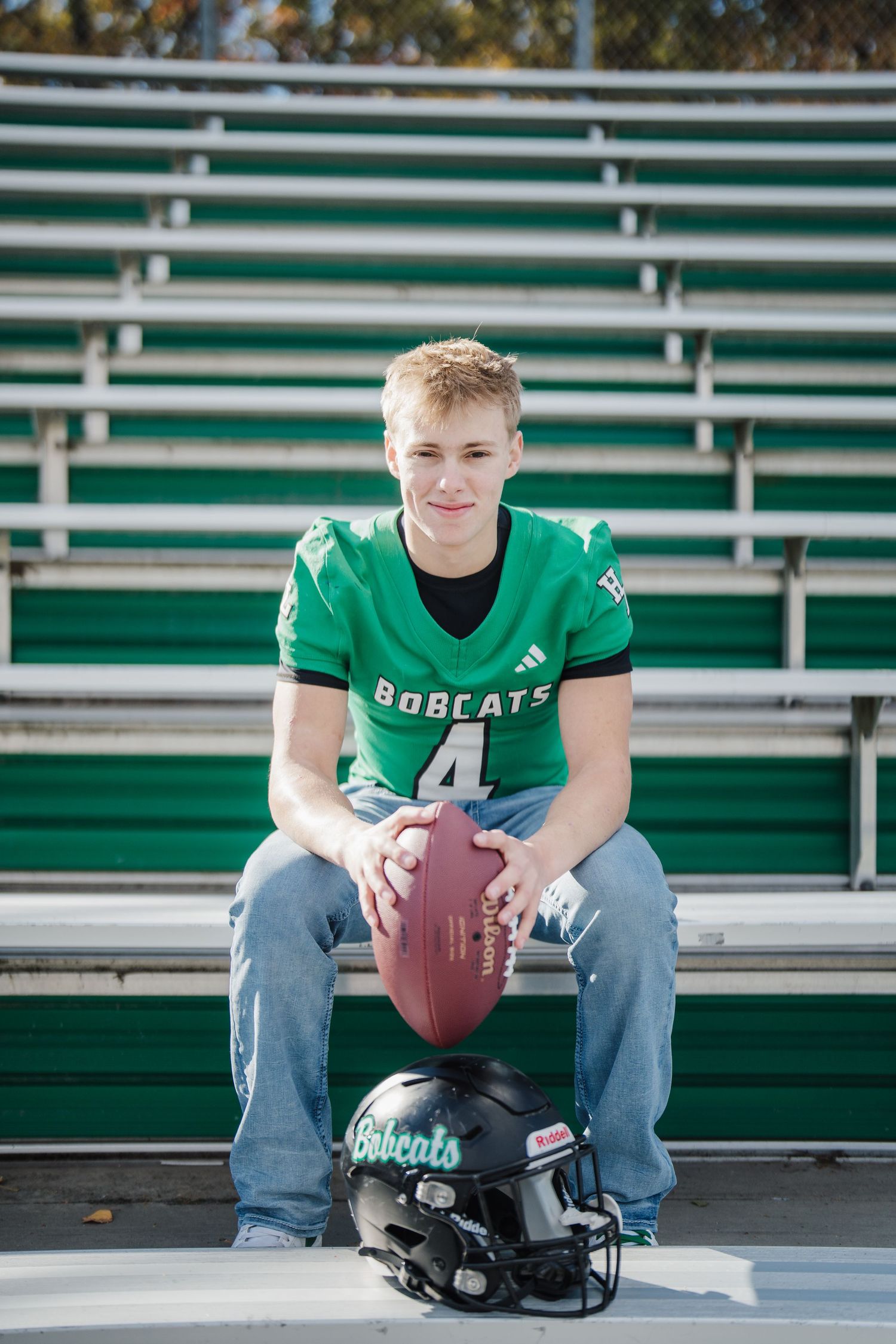 Player in green Bobcats jersey sits on stadium bleachers holding football with helmet at feet.