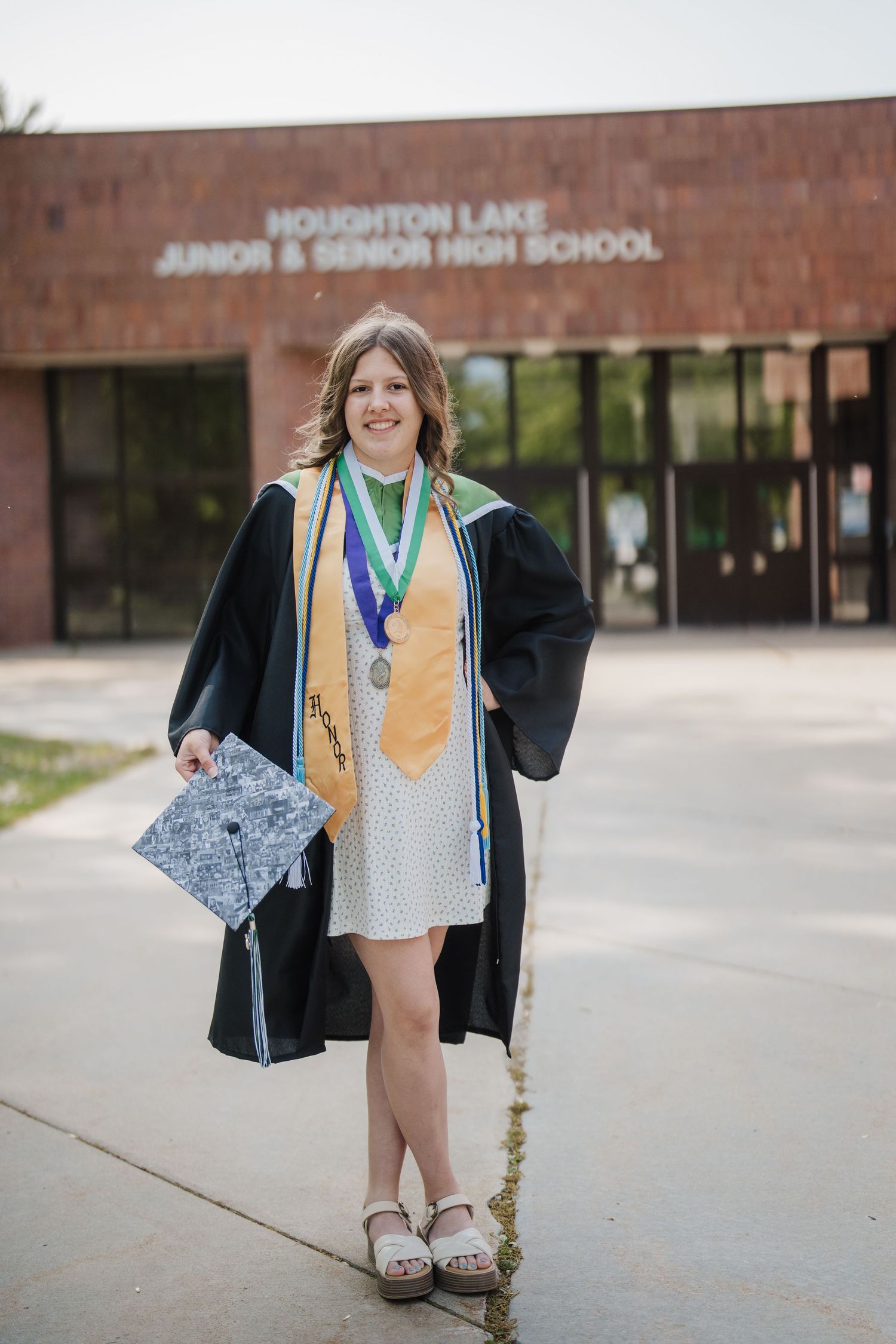 A graduate in cap and gown poses in front of Houghton Lake Junior High School entrance for photos.