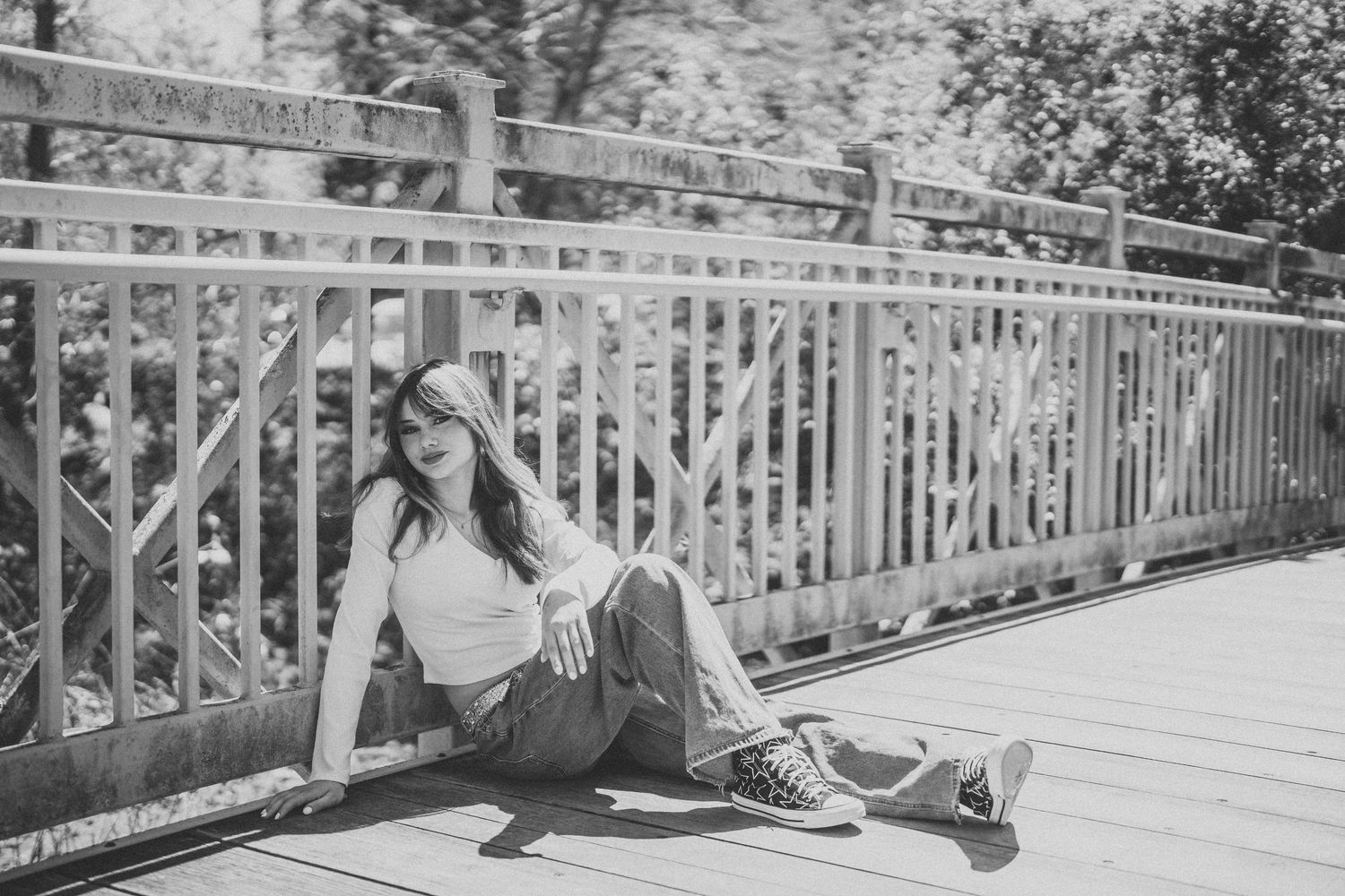 Black and white photo of someone sitting casually on a bridge walkway wearing a crop top and jeans on a sunny day.