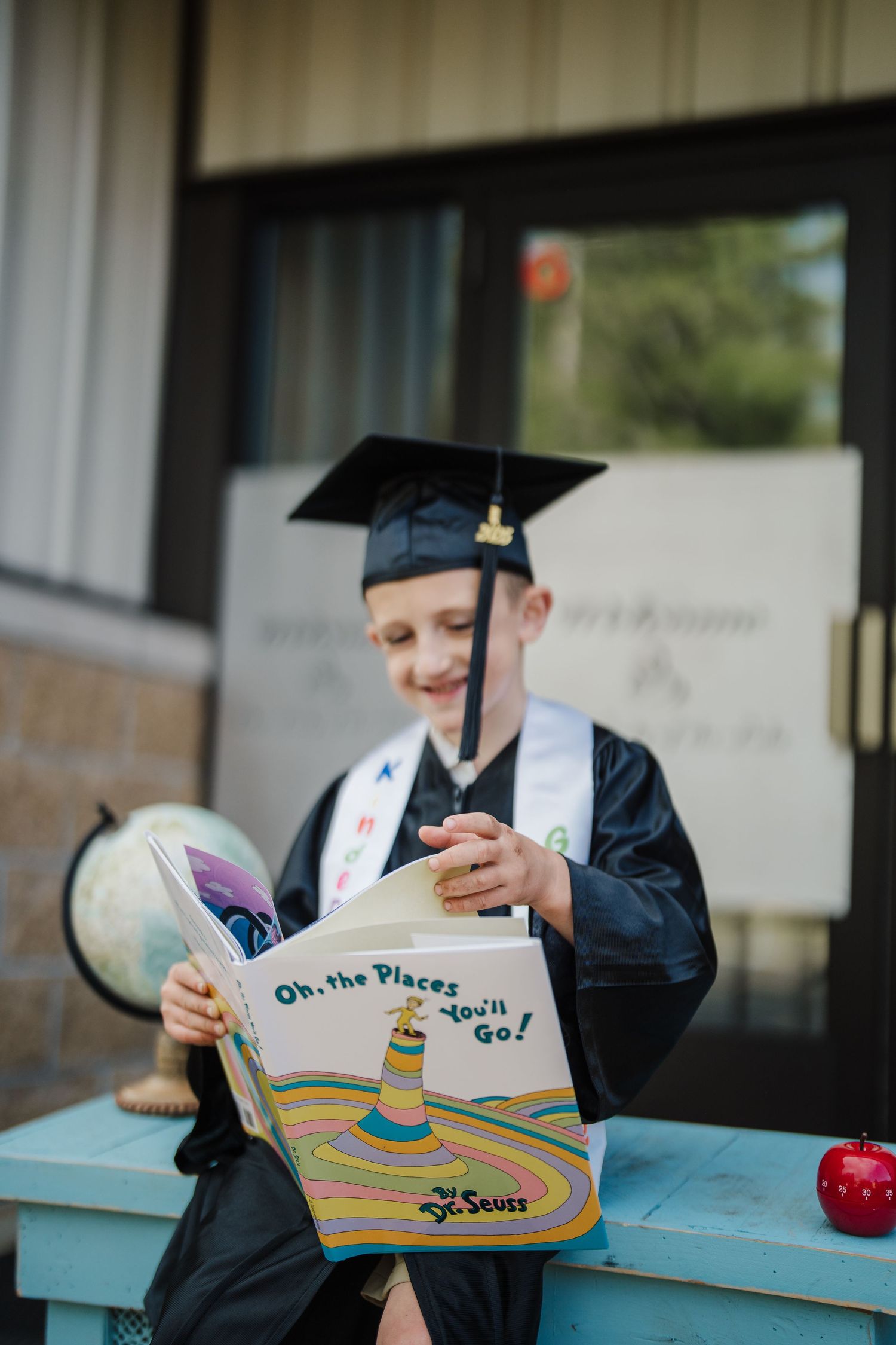 A graduate in cap and gown reads 'Oh The Places You'll Go' by Dr. Seuss while sitting at a blue table outdoors.