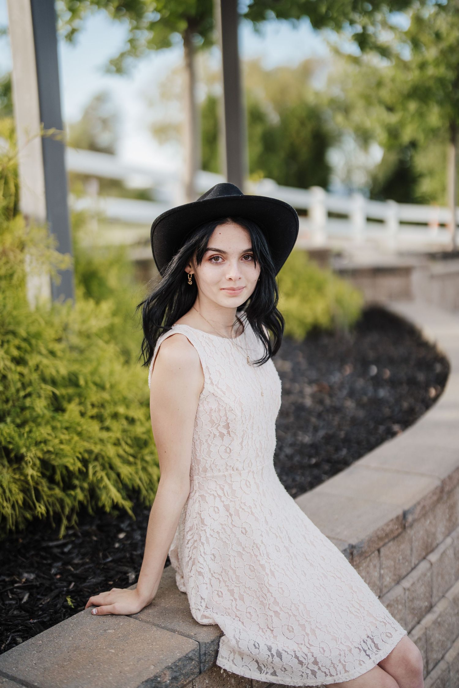 A person in a white lace dress and black sun hat poses on stone steps in an outdoor garden setting.