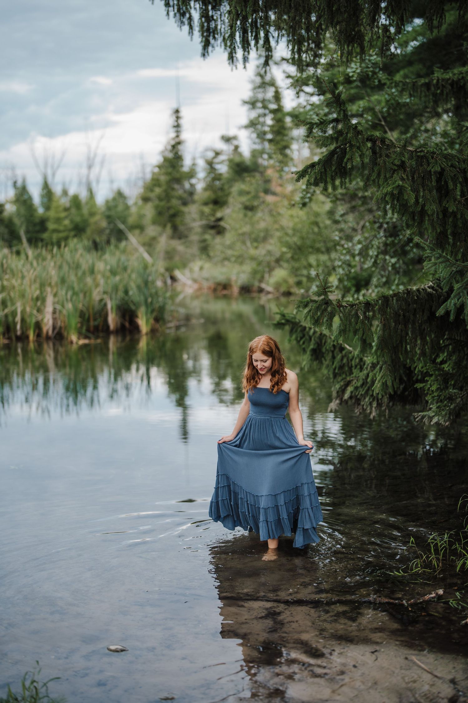 A blue dress flows gracefully near the water's edge surrounded by evergreen trees and reflective lake waters.