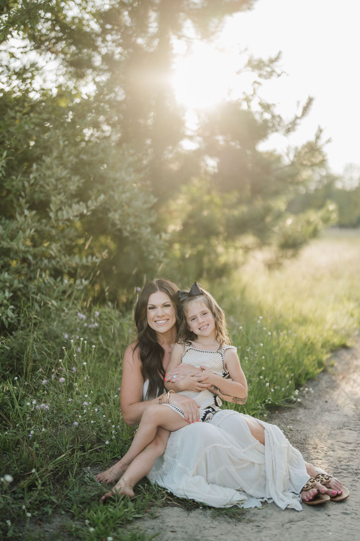 A parent and child sit together on a dirt path surrounded by tall grass during golden hour.