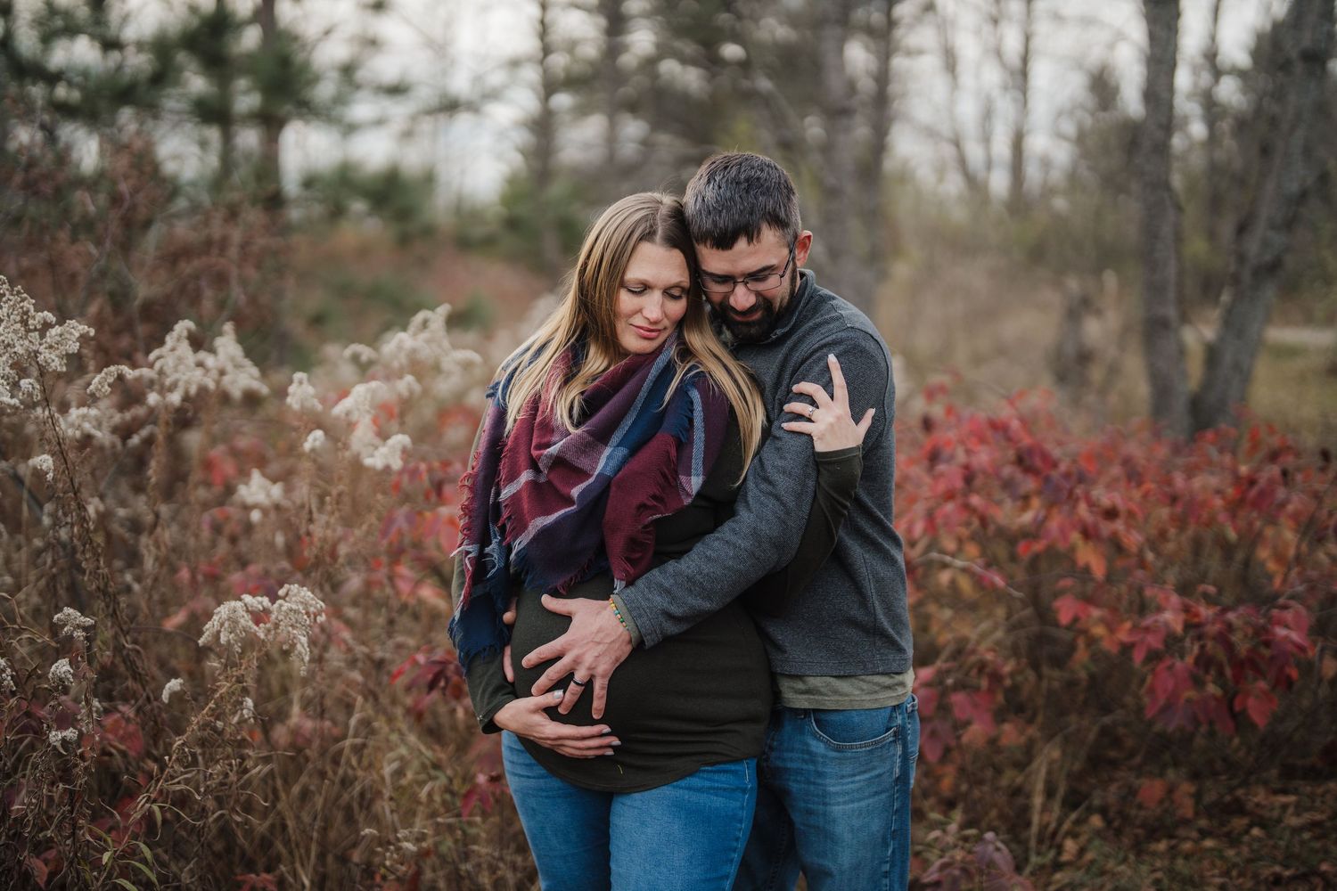 A couple embraces outdoors in autumn surrounded by red foliage and natural scenery during a romantic photoshoot.
