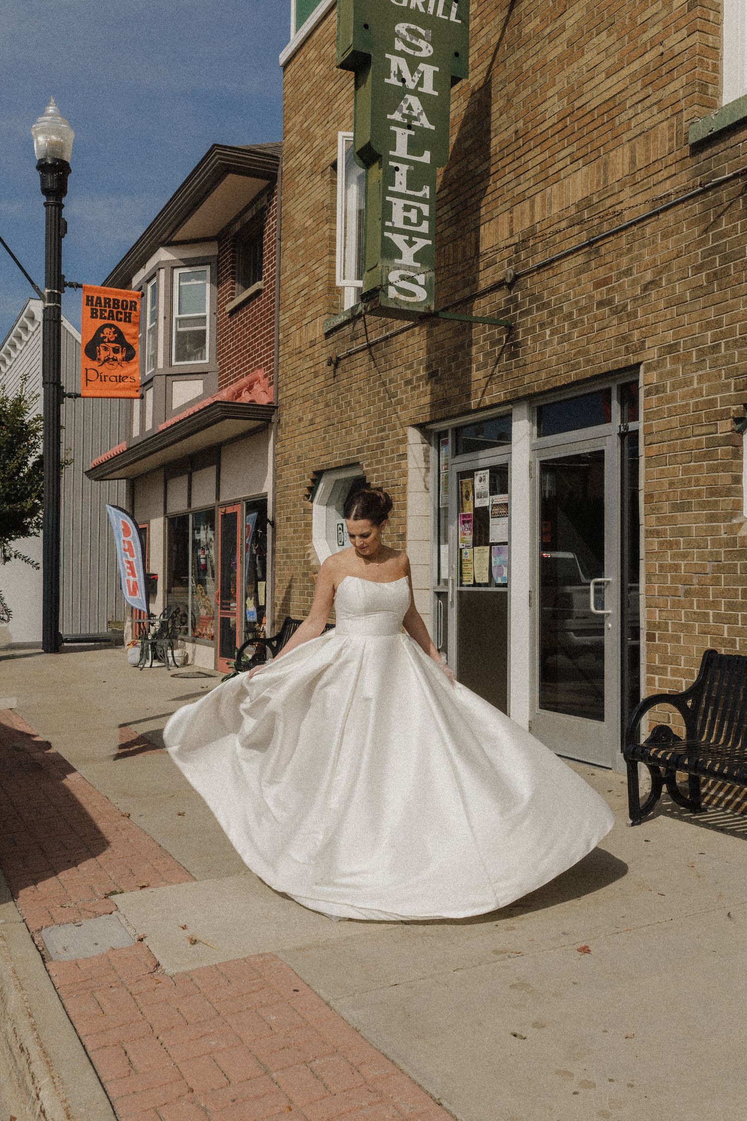 Bride in flowing white wedding dress twirling on brick-lined sidewalk outside storefronts.
