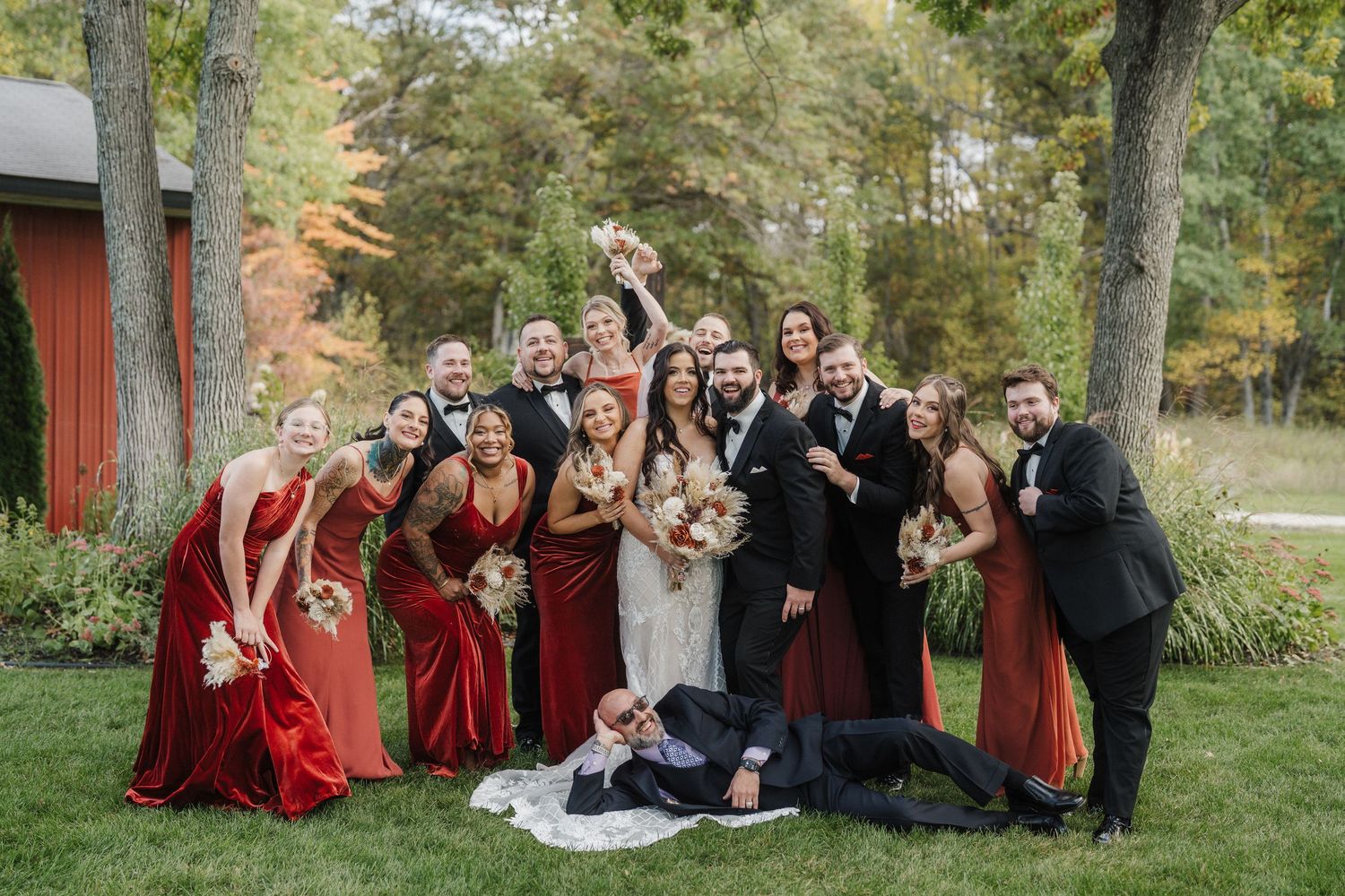 Wedding party in red dresses and black suits poses playfully on grass during outdoor autumn photoshoot.