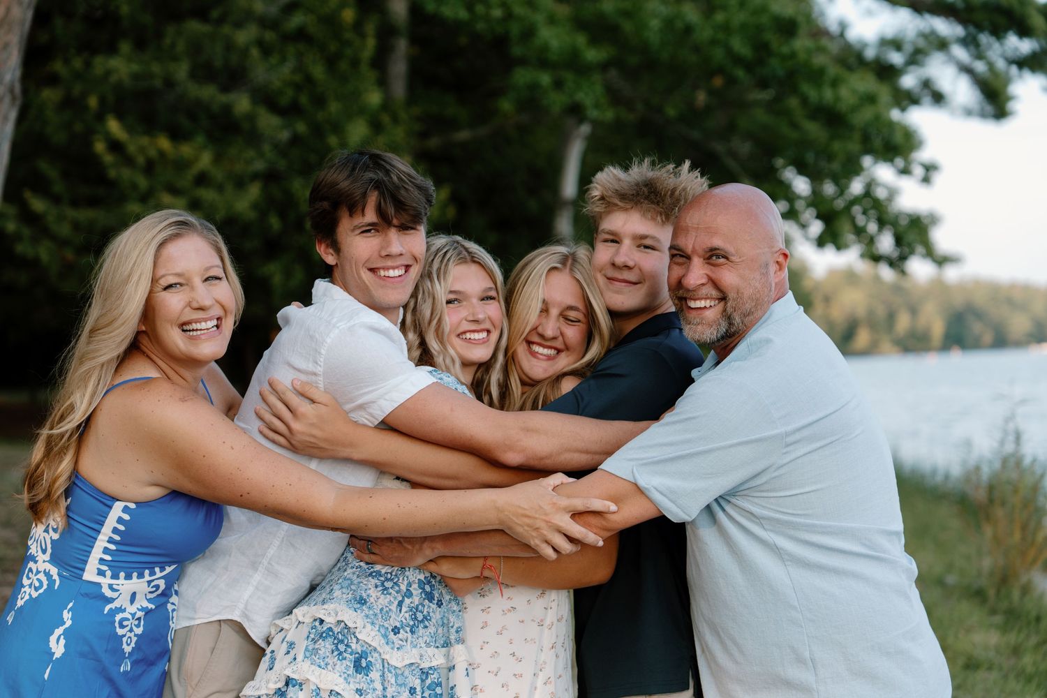 Happy family of six hugging outdoors near a lake surrounded by trees during a warm summer evening.
