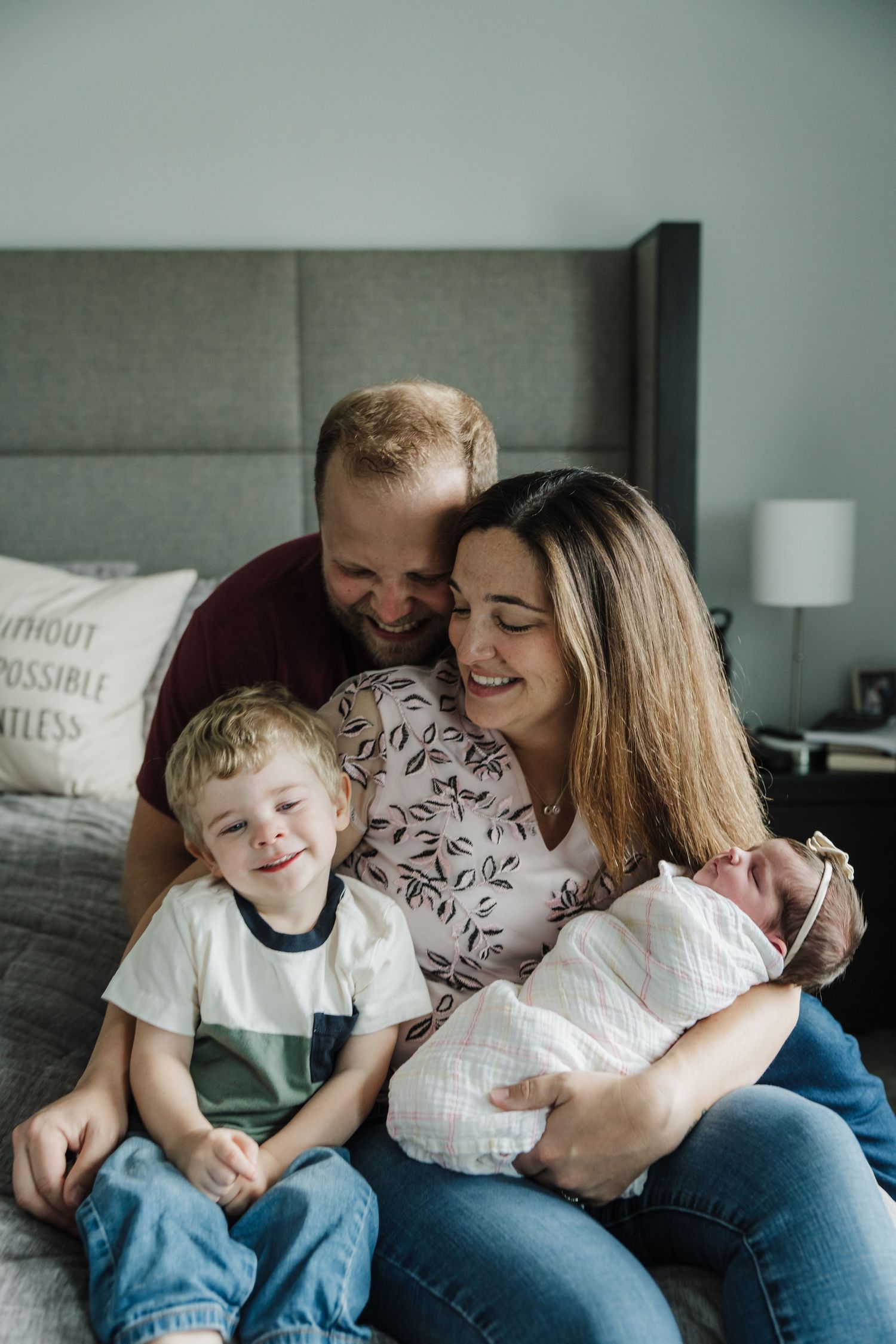 Happy family of four sitting on a bed, parents smiling at newborn baby while young blond toddler boy grins at camera.