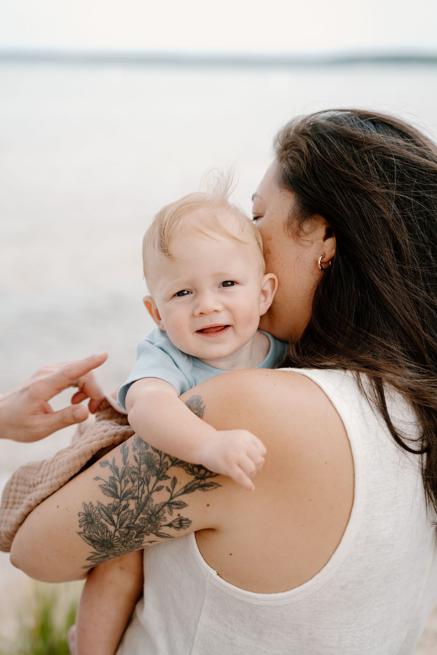 A tattooed mother kisses her smiling baby outdoors near a lake, holding the infant in her arms.