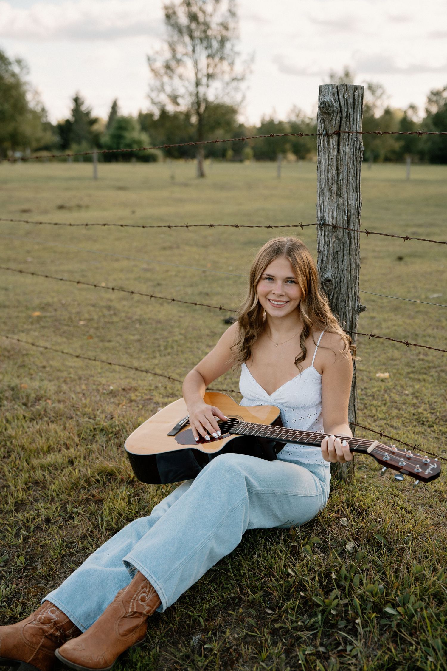 Young woman smiling while playing acoustic guitar outdoors, sitting by a rustic fence post in a green field wearing cowboy boots.