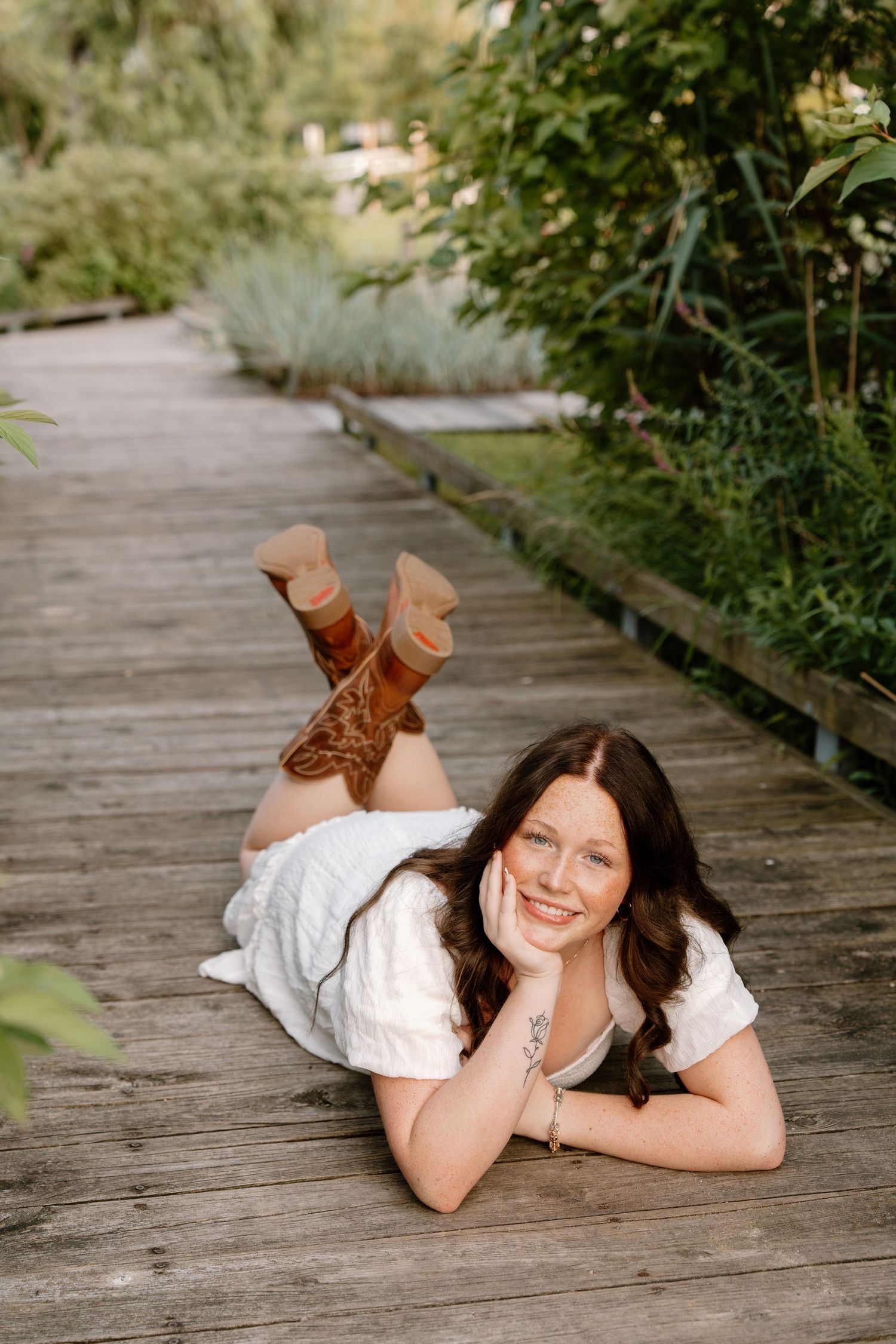 Young woman in white dress and cowboy boots lying on wooden boardwalk path surrounded by lush green garden.