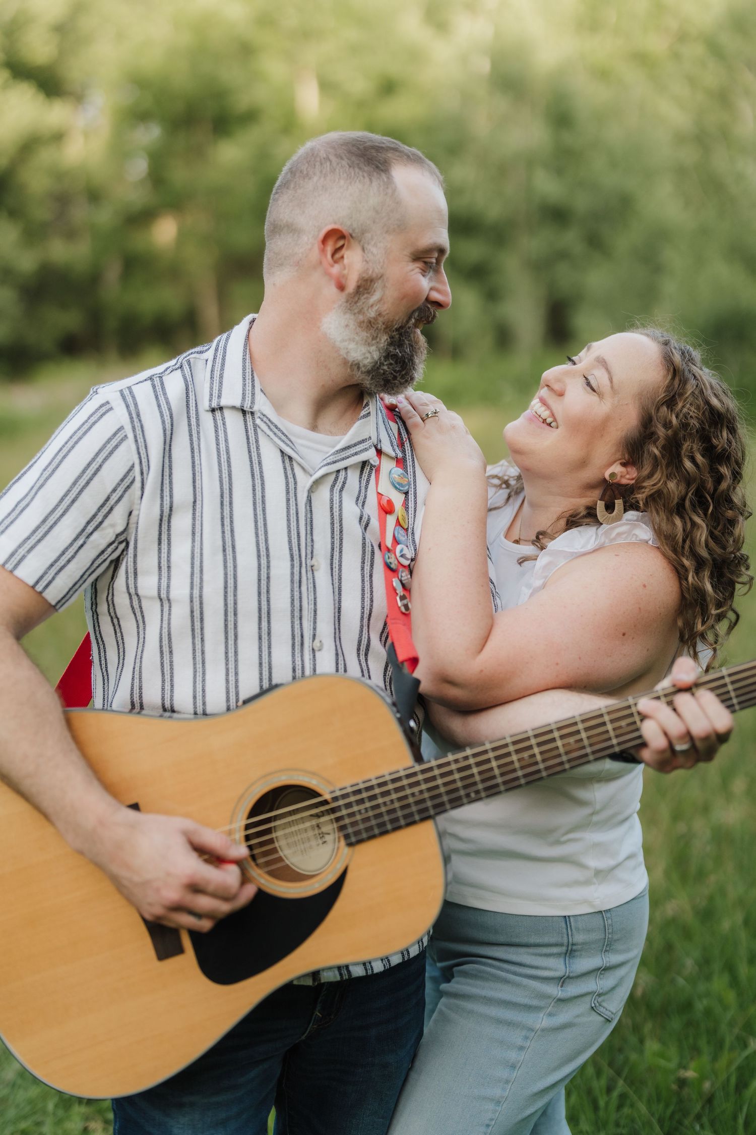 A bearded man plays acoustic guitar outdoors while a curly-haired woman smiles and leans on his shoulder.