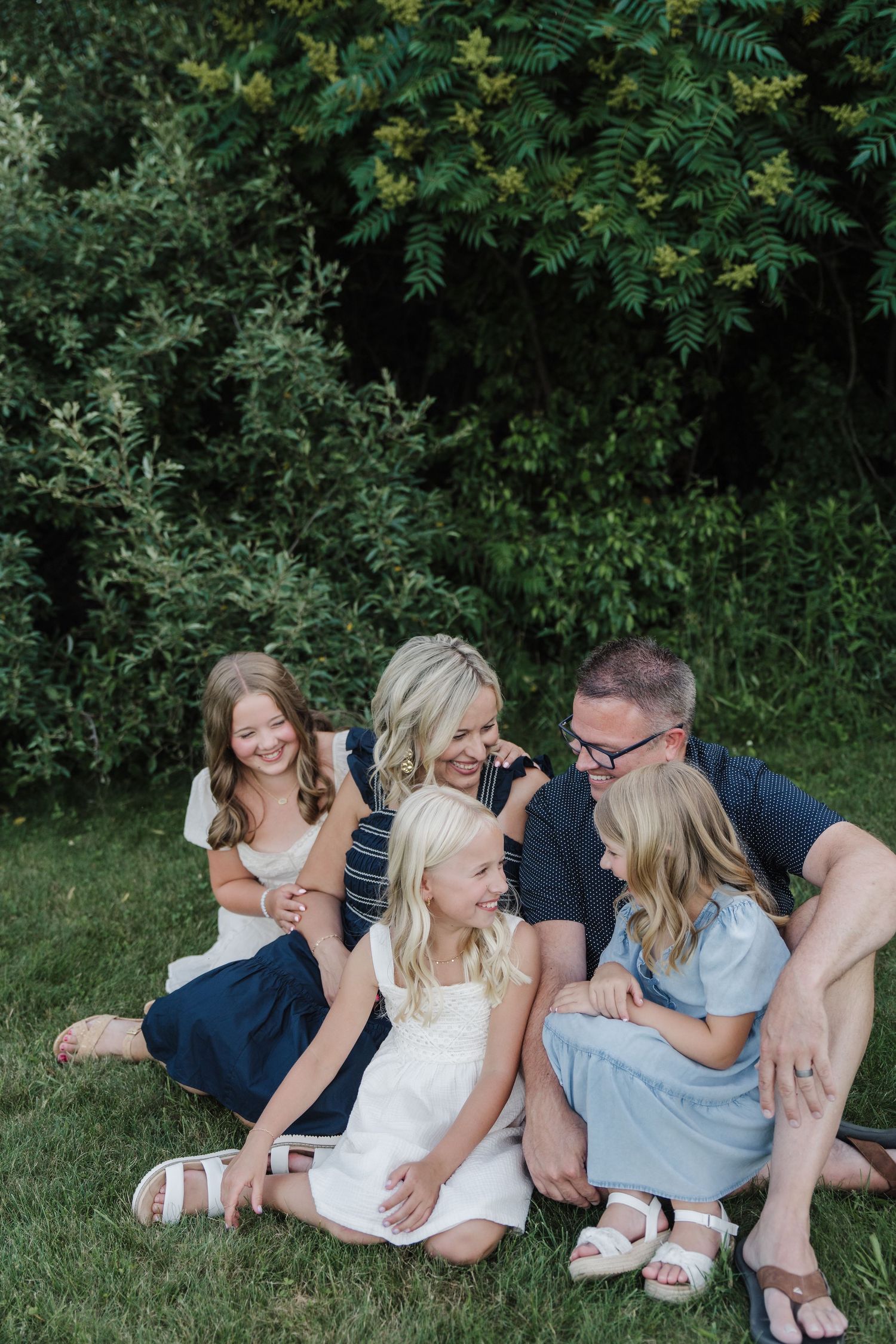 Happy family of five sitting together on green grass, laughing and embracing outdoors near lush bushes.