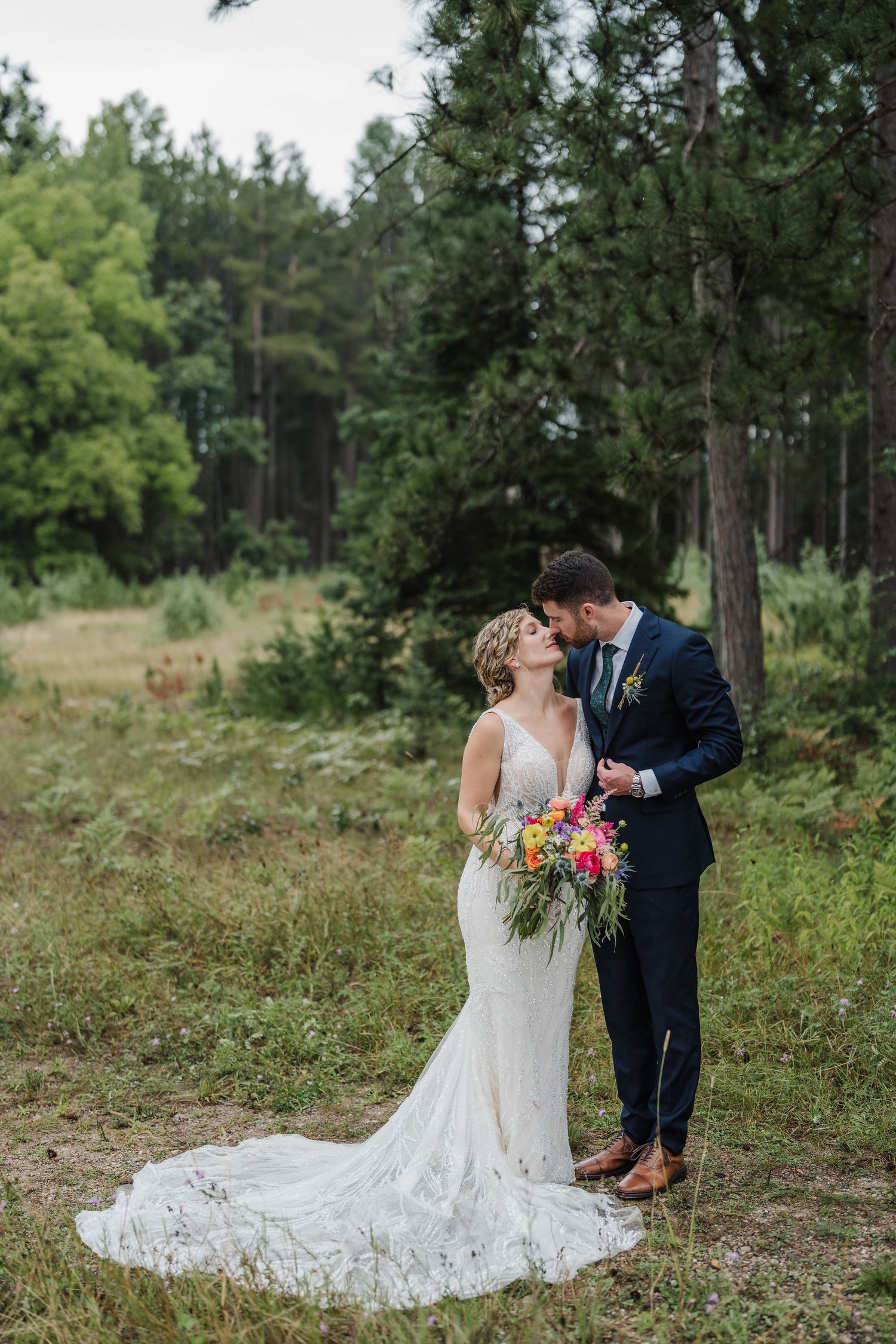 A wedding couple poses in a romantic forest setting with a colorful bouquet.
