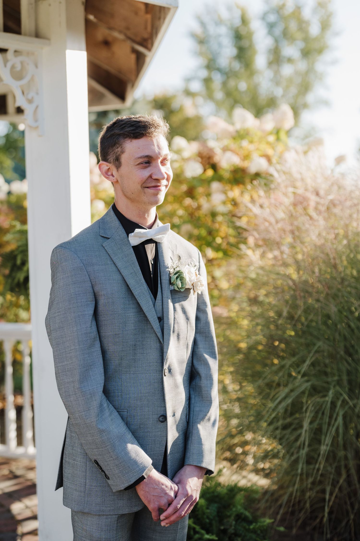 Groom in light grey suit and bowtie smiles while standing on white porch.