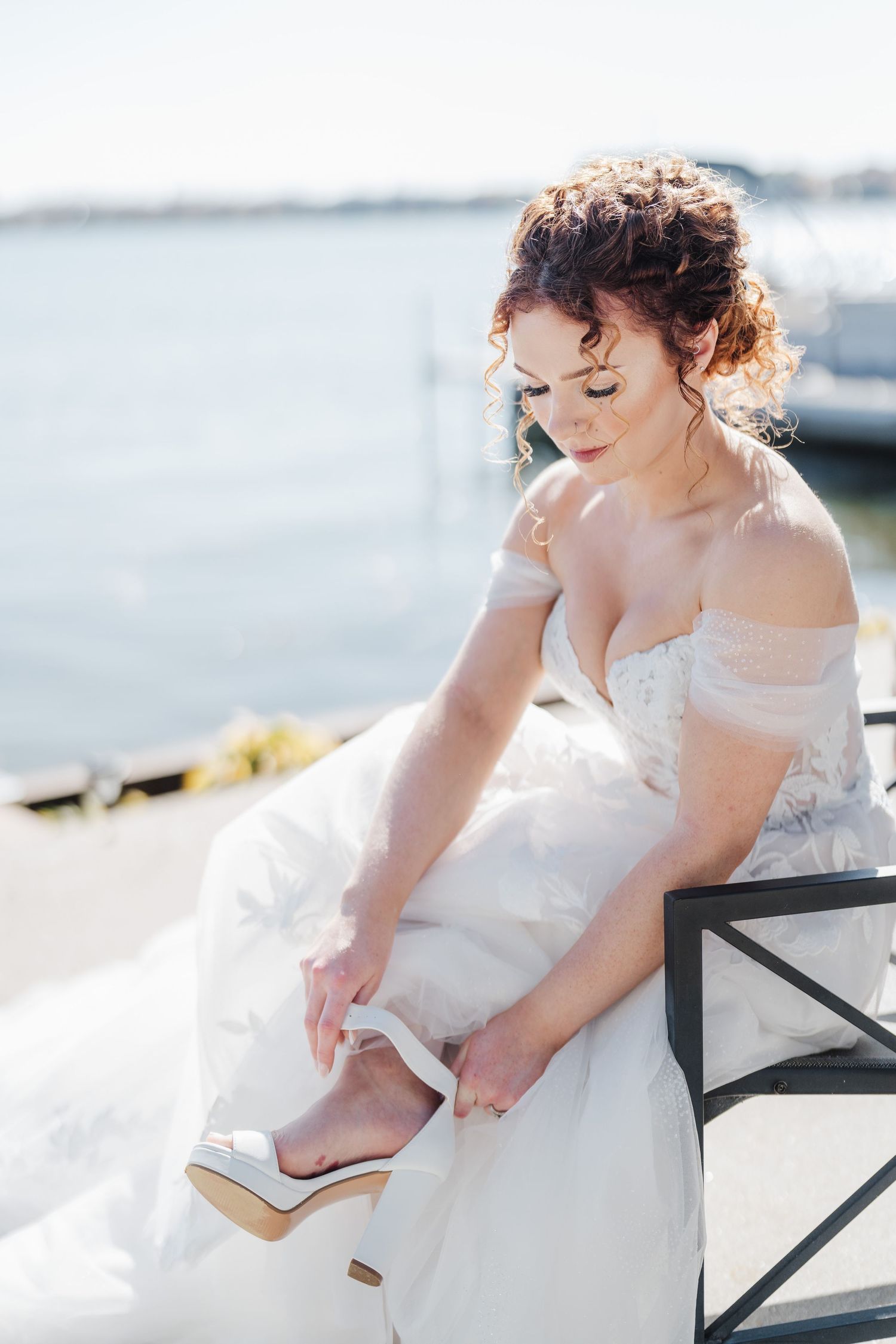 Person in off-shoulder wedding dress adjusts their shoe while sitting in bright natural light.