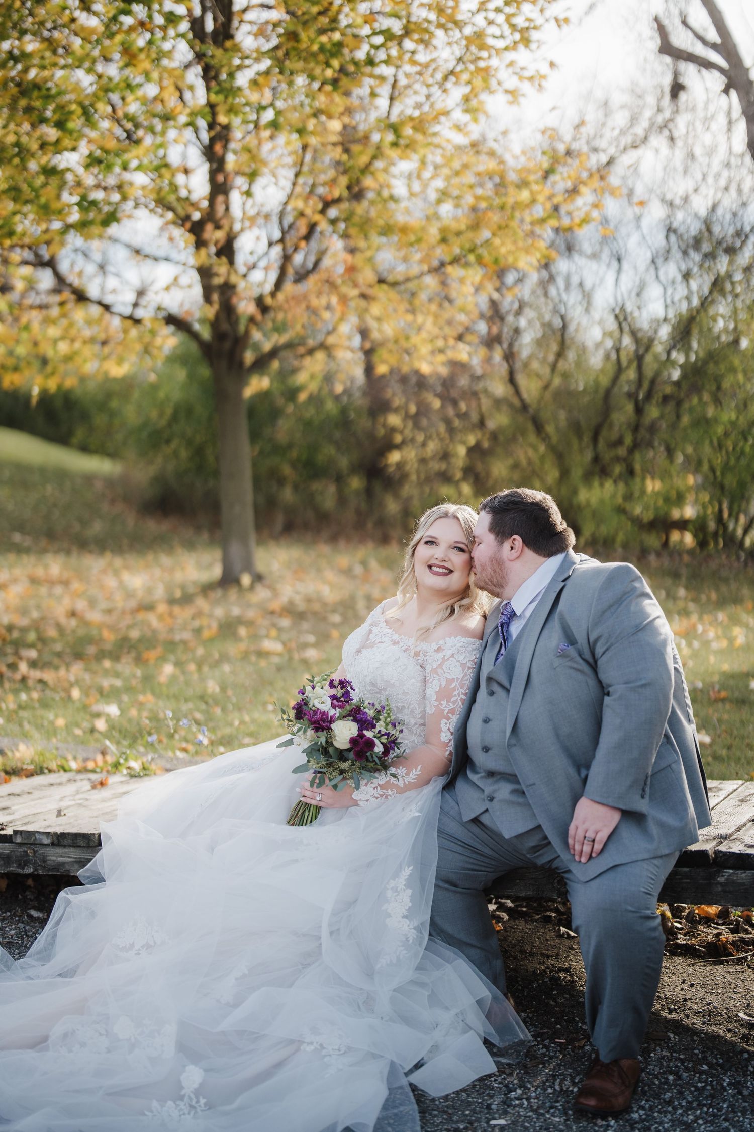 Newlyweds share intimate moment while seated on bench surrounded by autumn leaves.