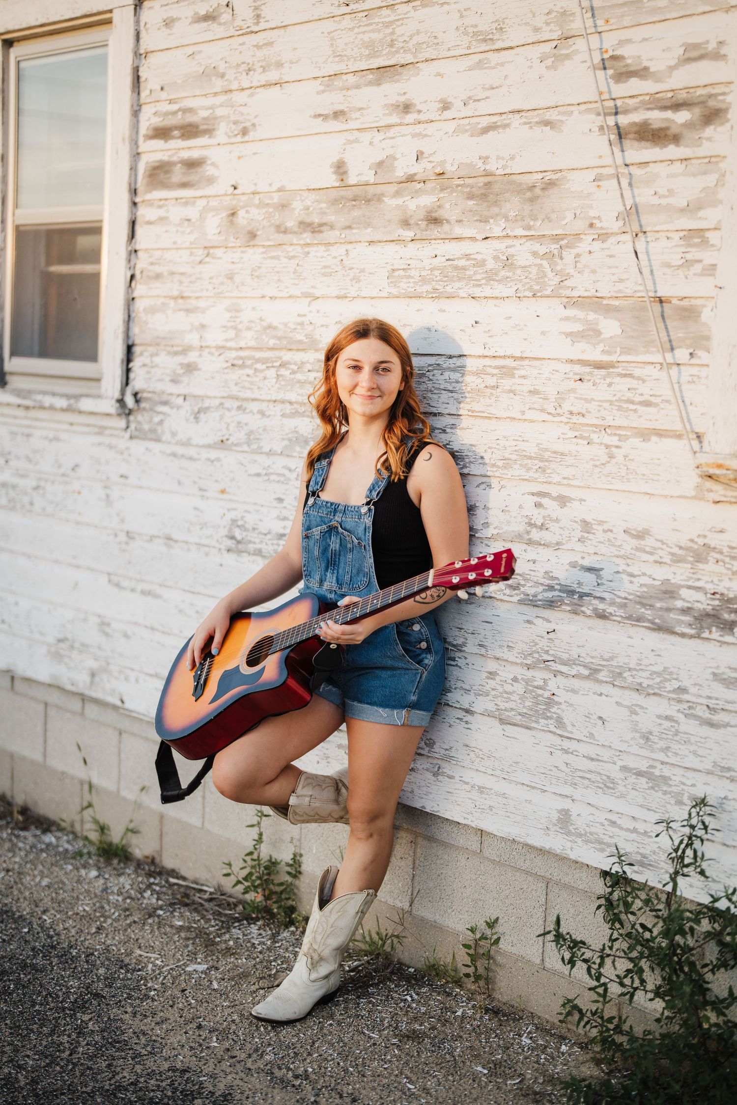 A musician in denim overalls sits against a weathered white wall playing an acoustic guitar.