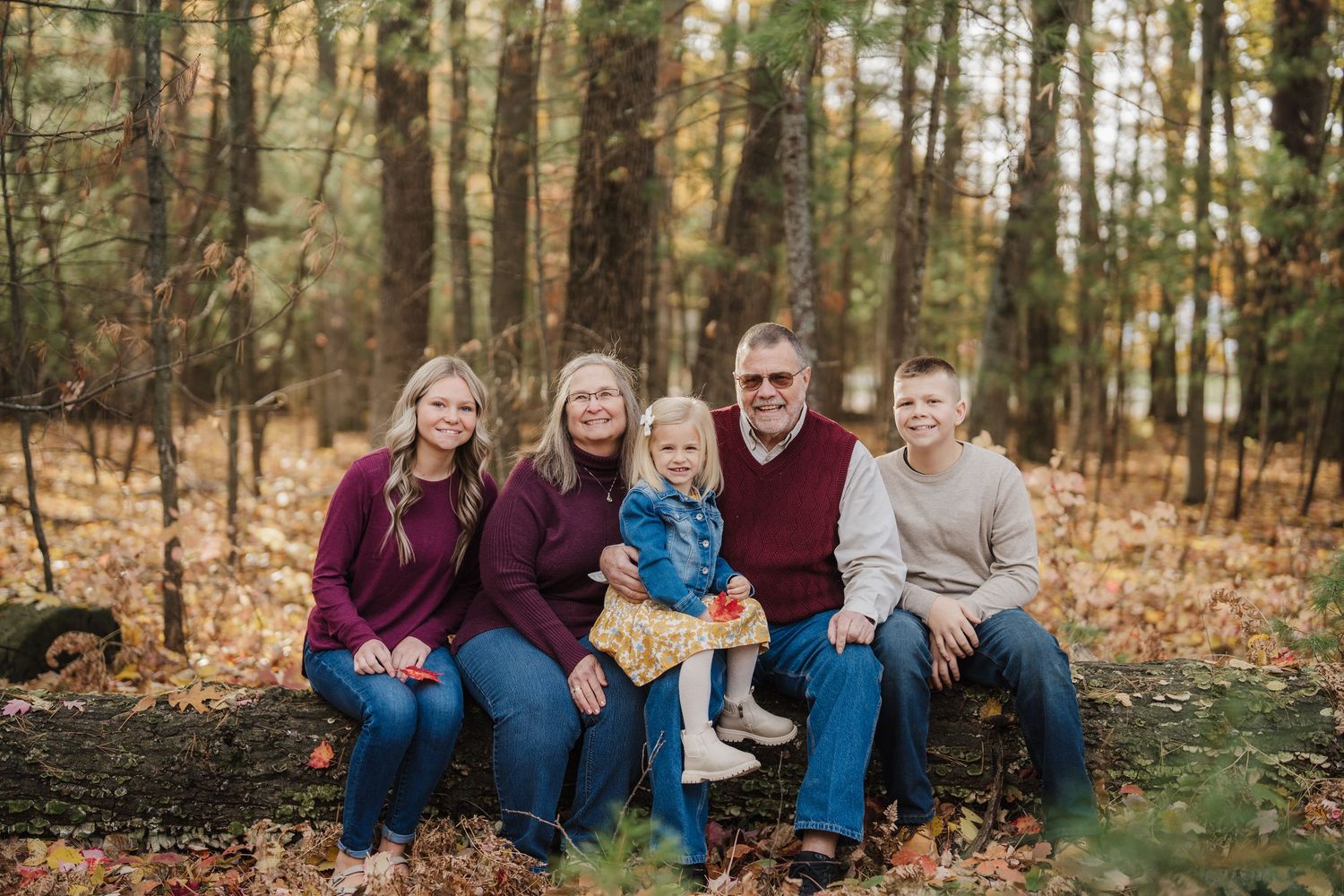 Family members sit together and laugh while posing for outdoor autumn portraits in a wooded setting.