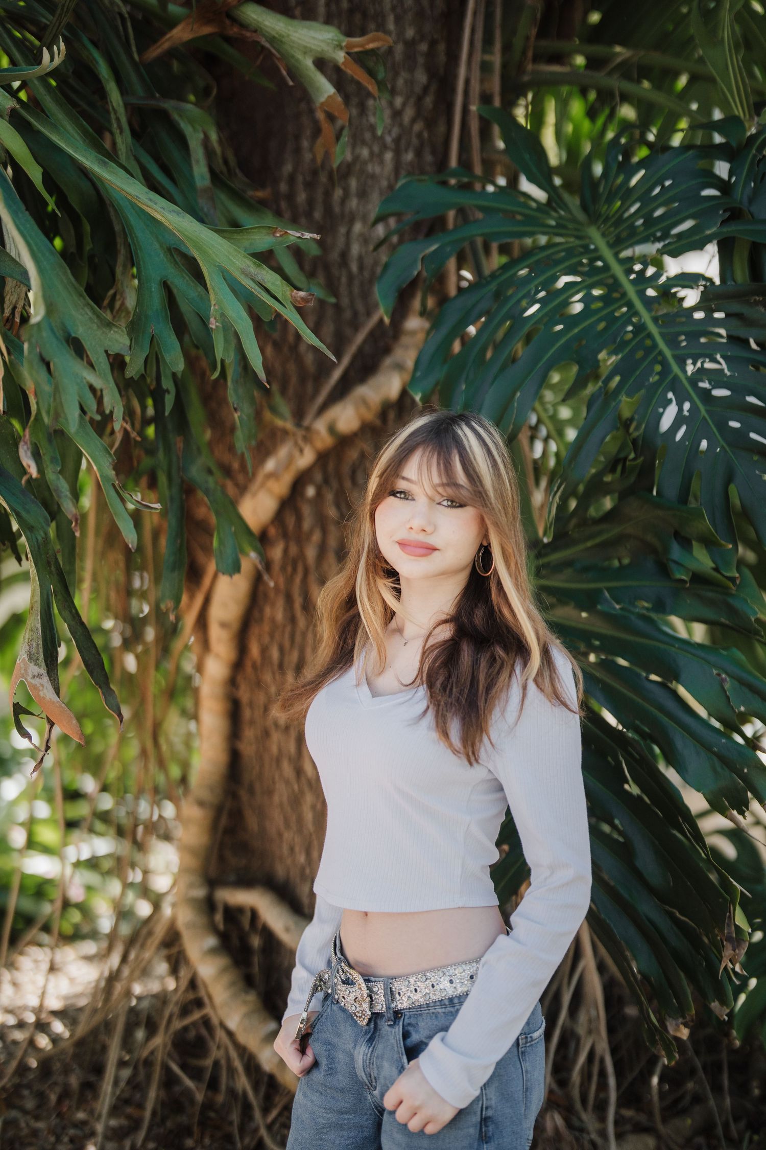 A woman in a light gray crop top and jeans poses outdoors against lush tropical foliage and trees.
