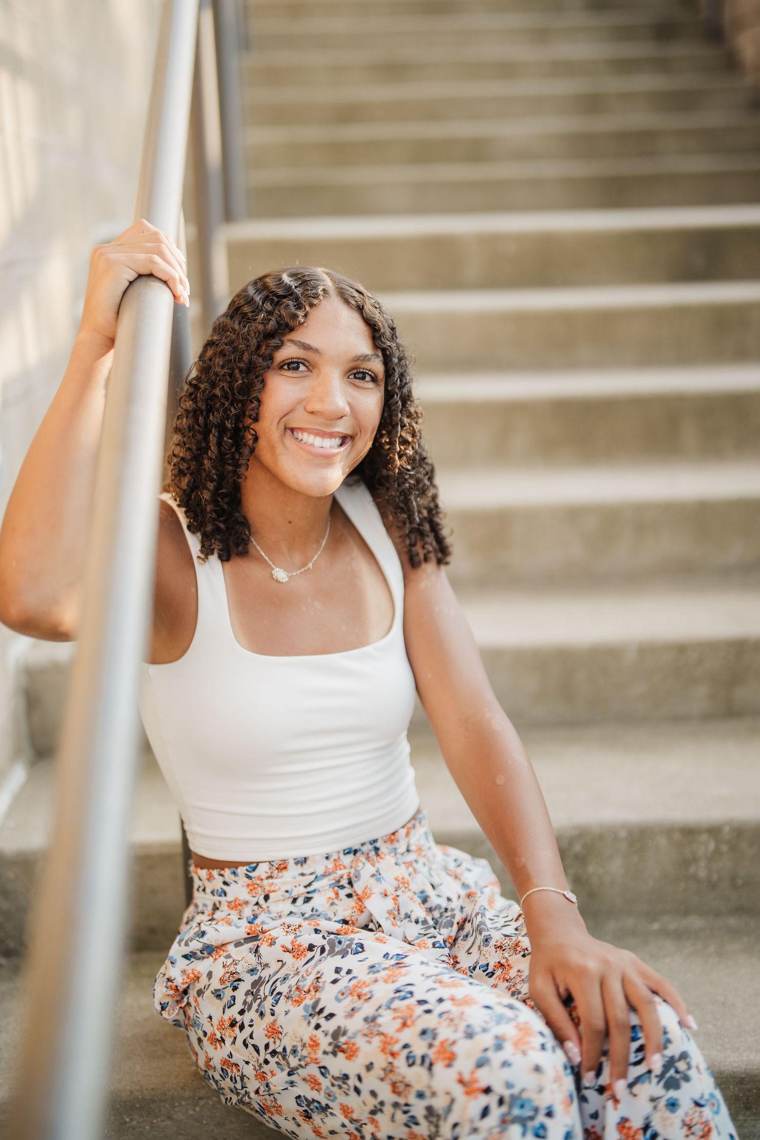 A woman with curly hair sits on concrete steps wearing a white tank top and floral pants on a sunny day.