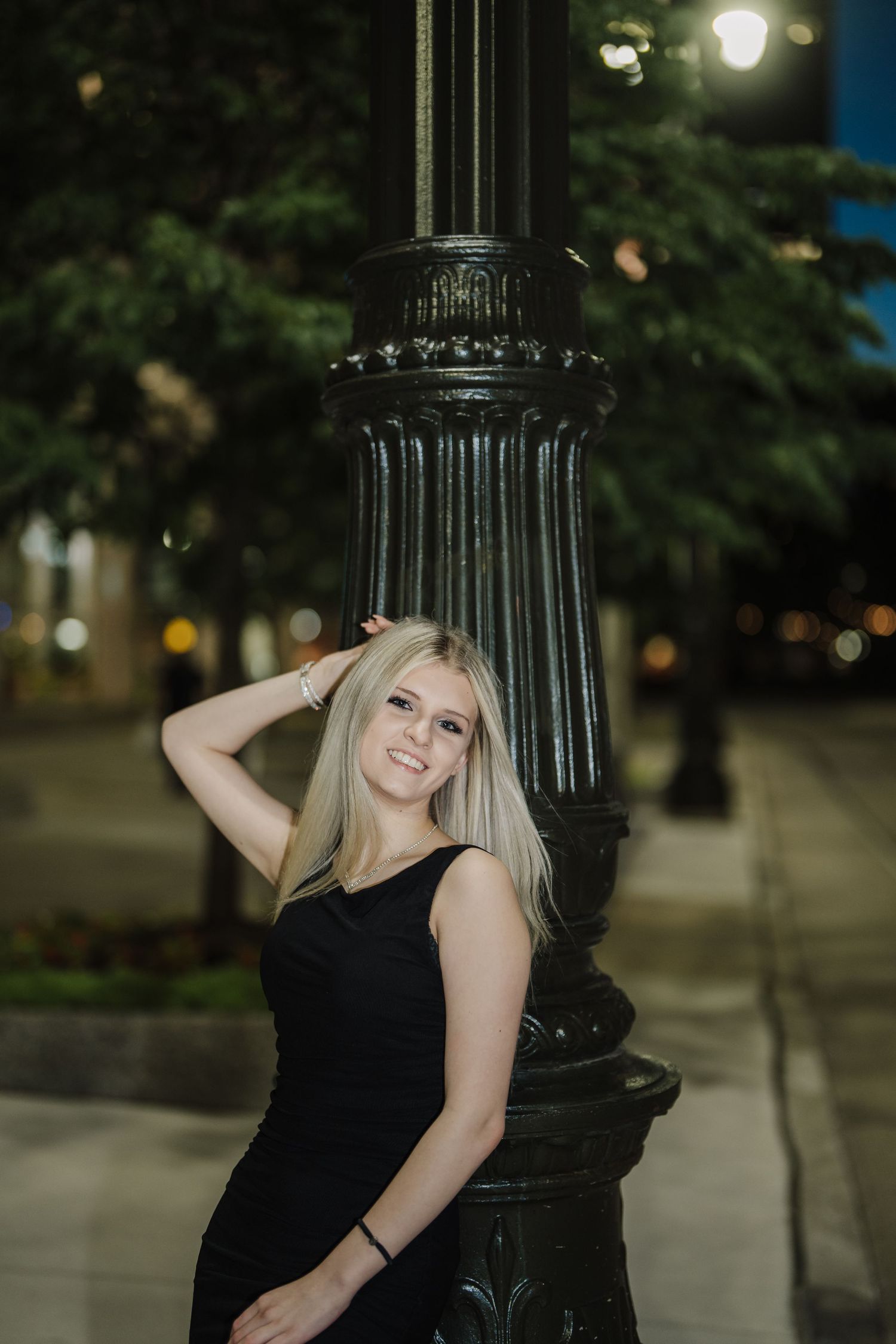 A person in a black dress poses next to a decorative lamp post in an outdoor evening setting.