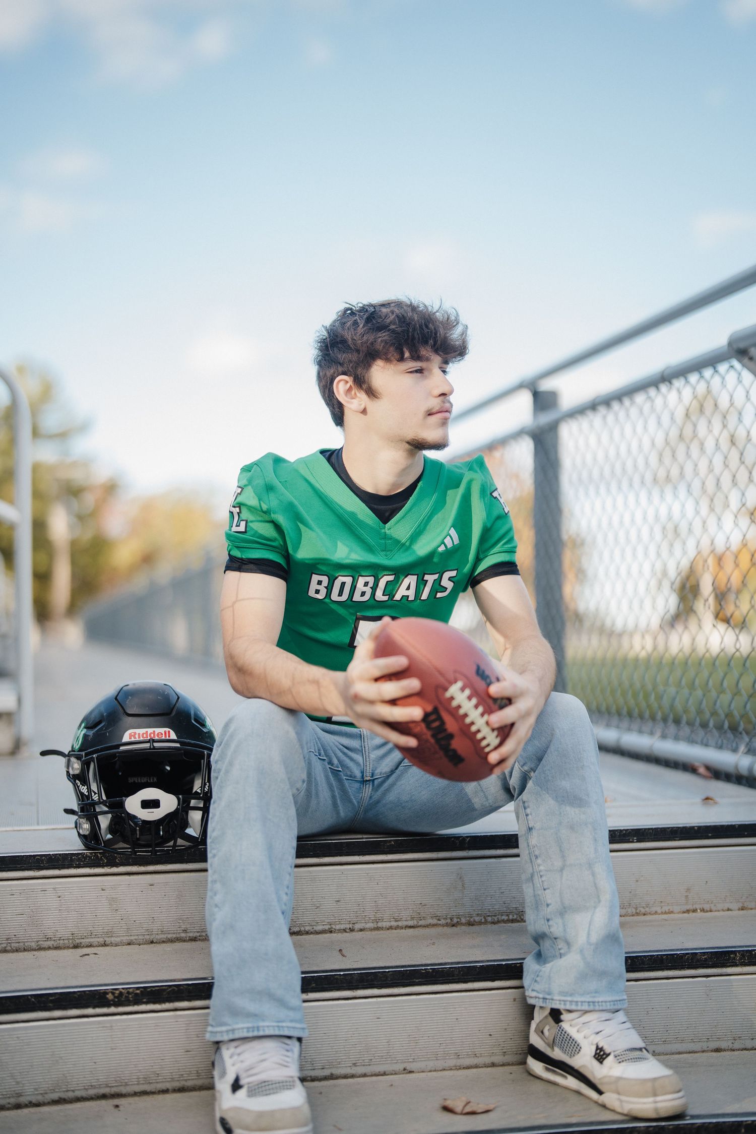 A football player in a green Bobcats jersey sits on bleachers holding a football.