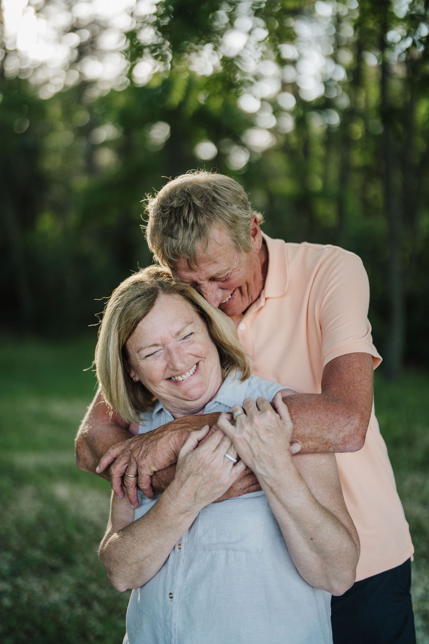Two people share a joyful embrace outdoors in a natural setting with trees in the background.