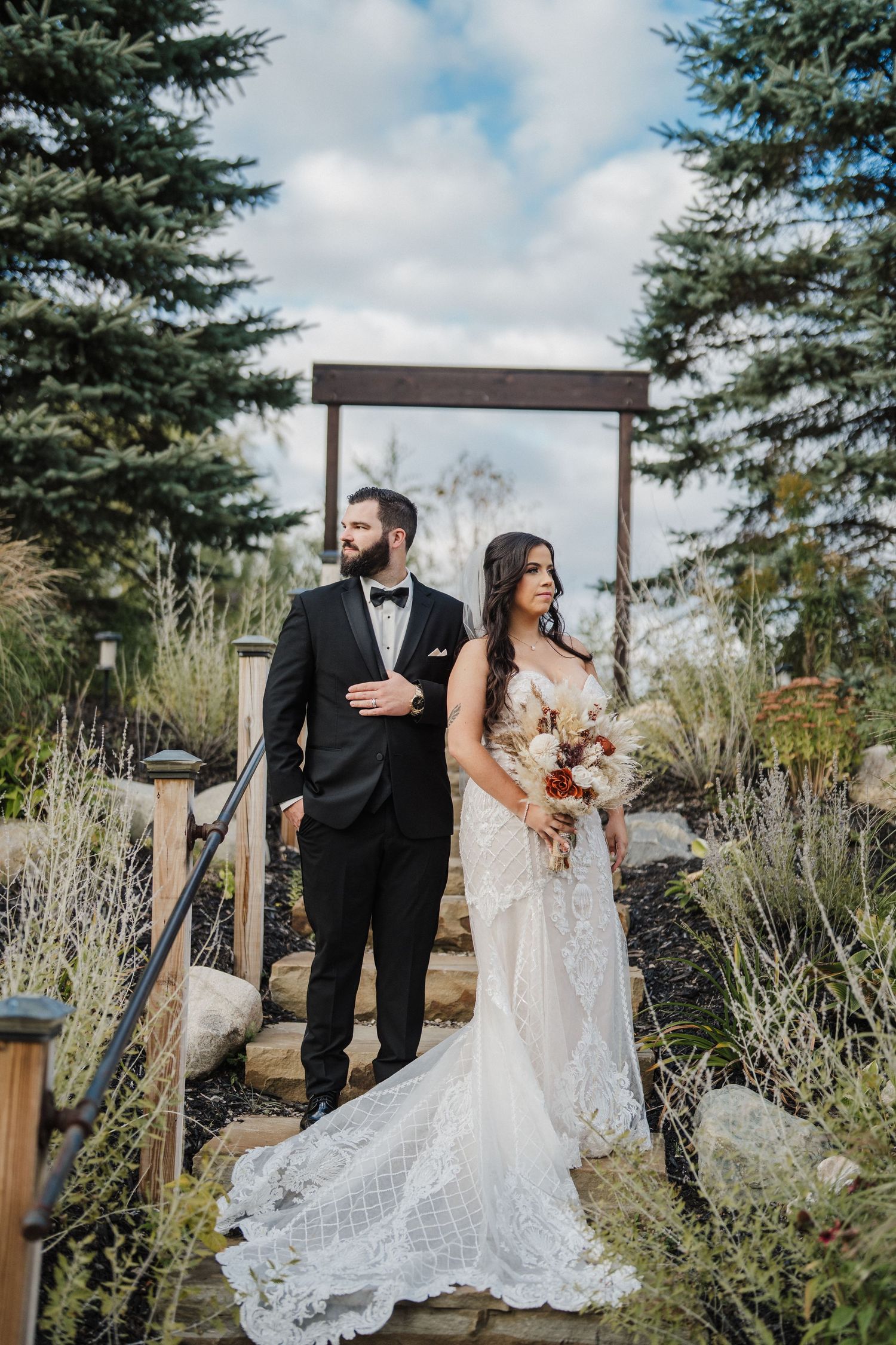 A wedding couple poses in a garden setting with evergreen trees and ornamental grasses creating a natural backdrop.