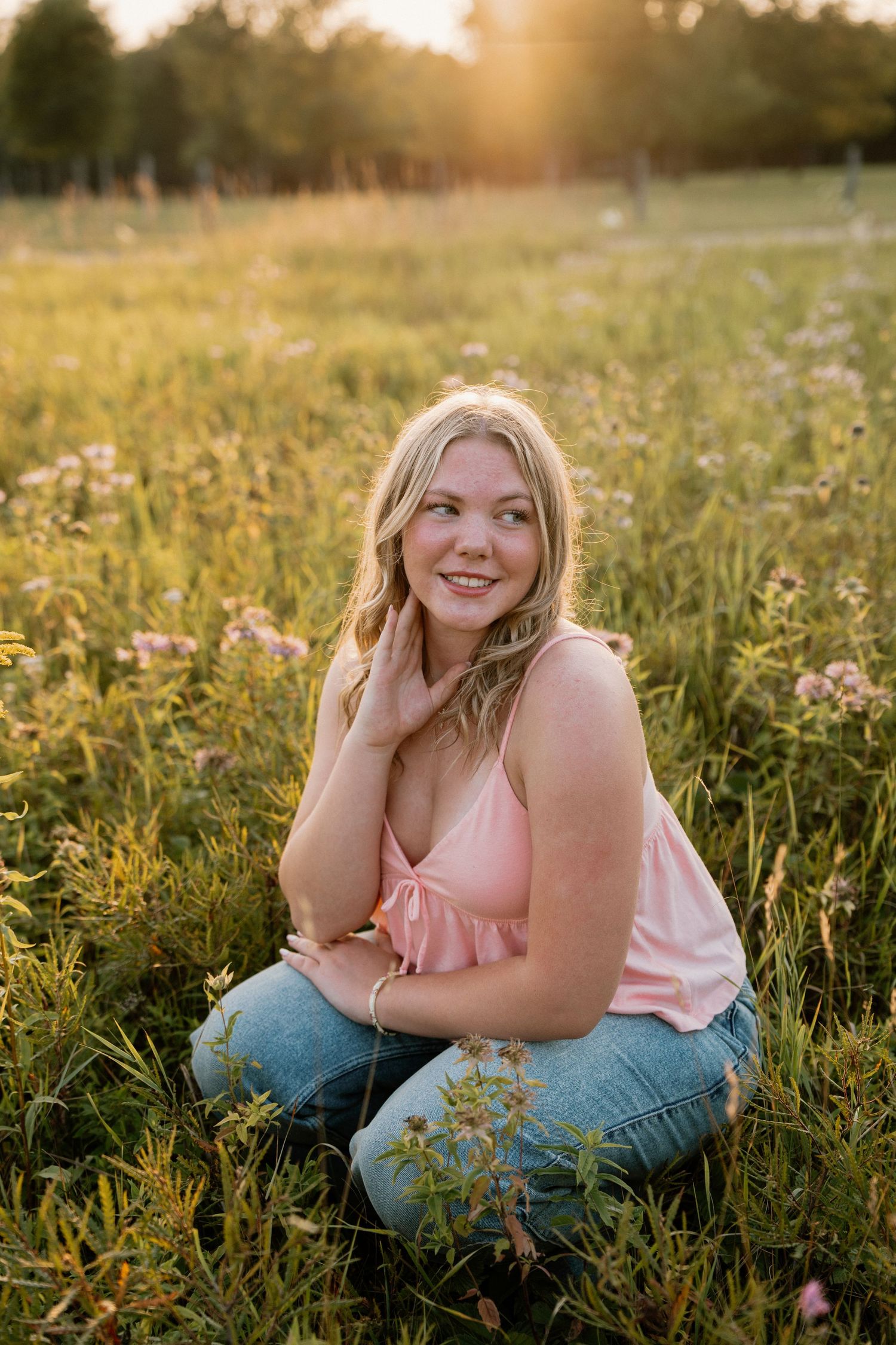 Young blonde woman in pink top and jeans sitting in a wildflower field during golden hour sunset.