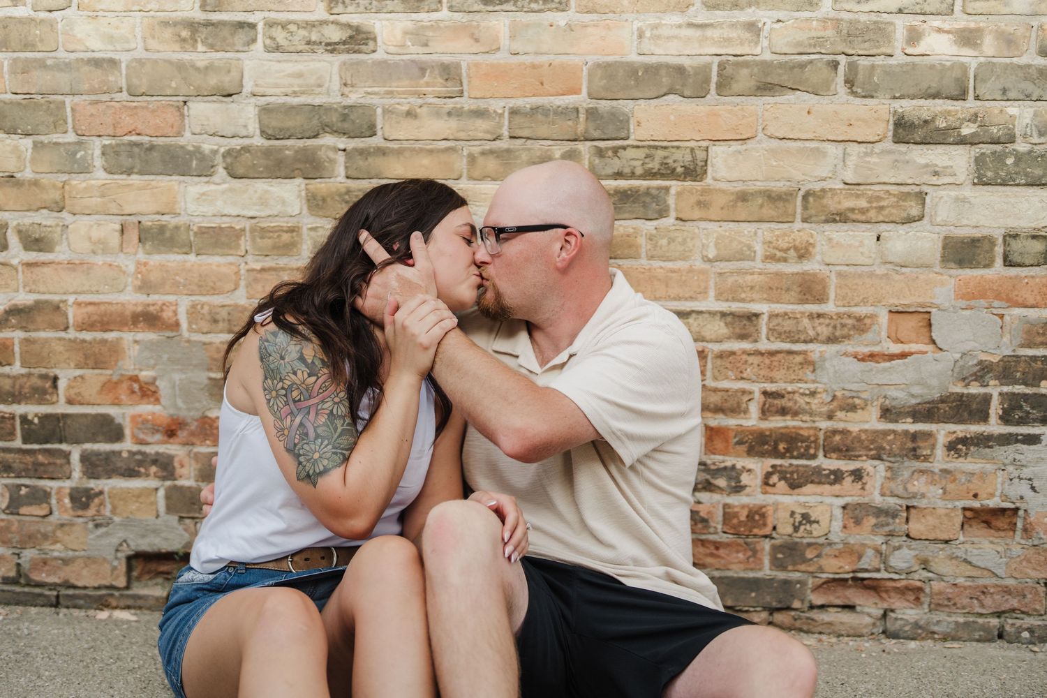 Couple sharing a romantic kiss while sitting against a rustic brick wall outdoors.