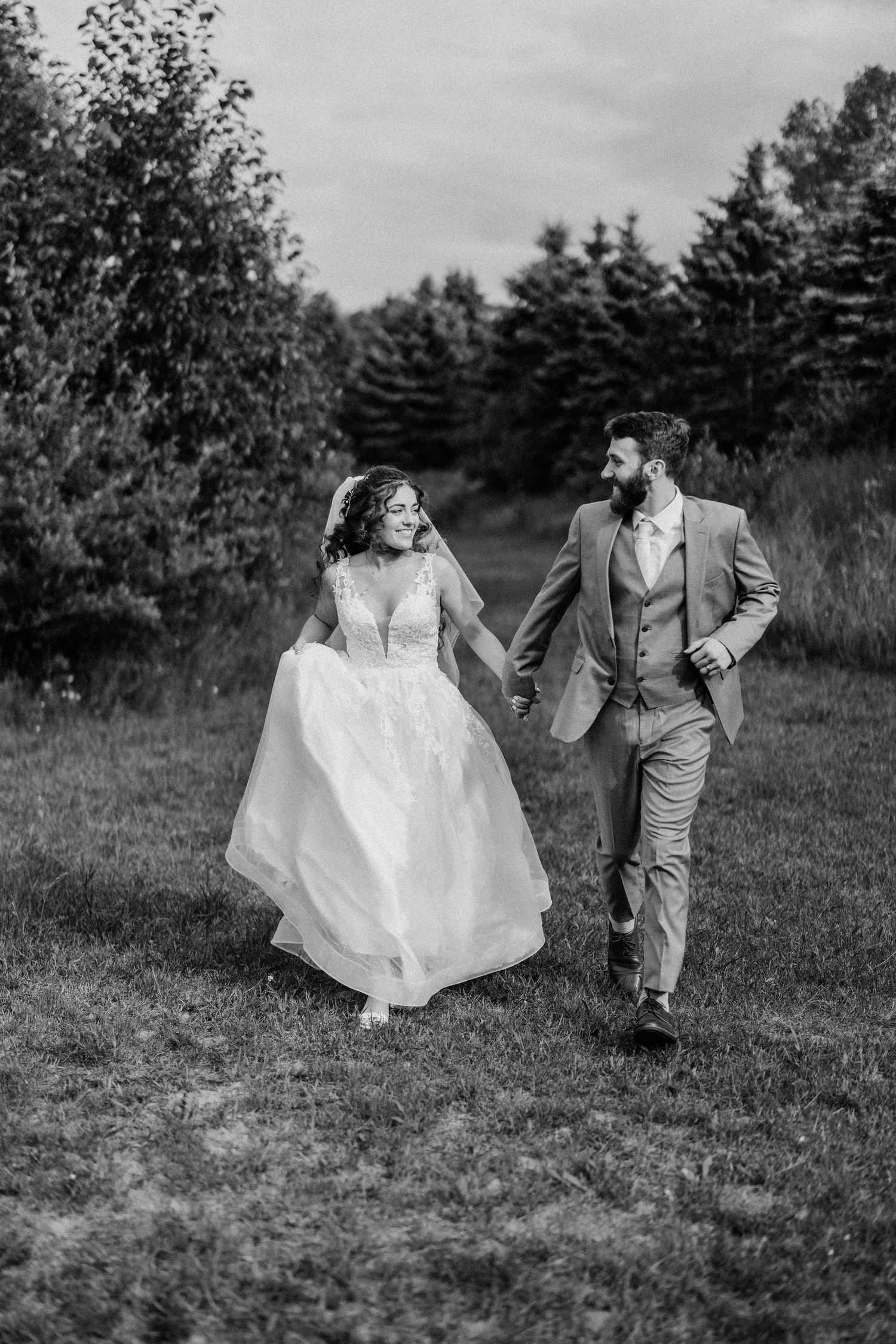 Black and white photo of newlyweds running together in a field with pine trees.