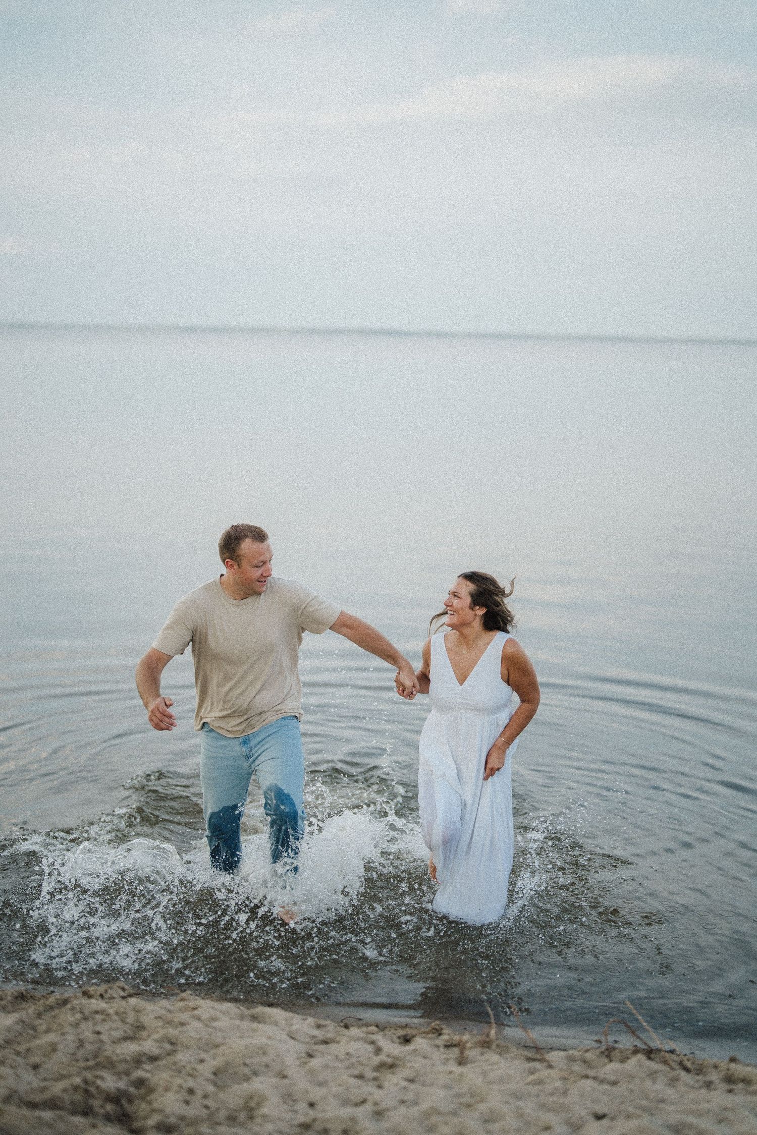 A couple splashes and plays together in shallow ocean water during a romantic beach photoshoot.