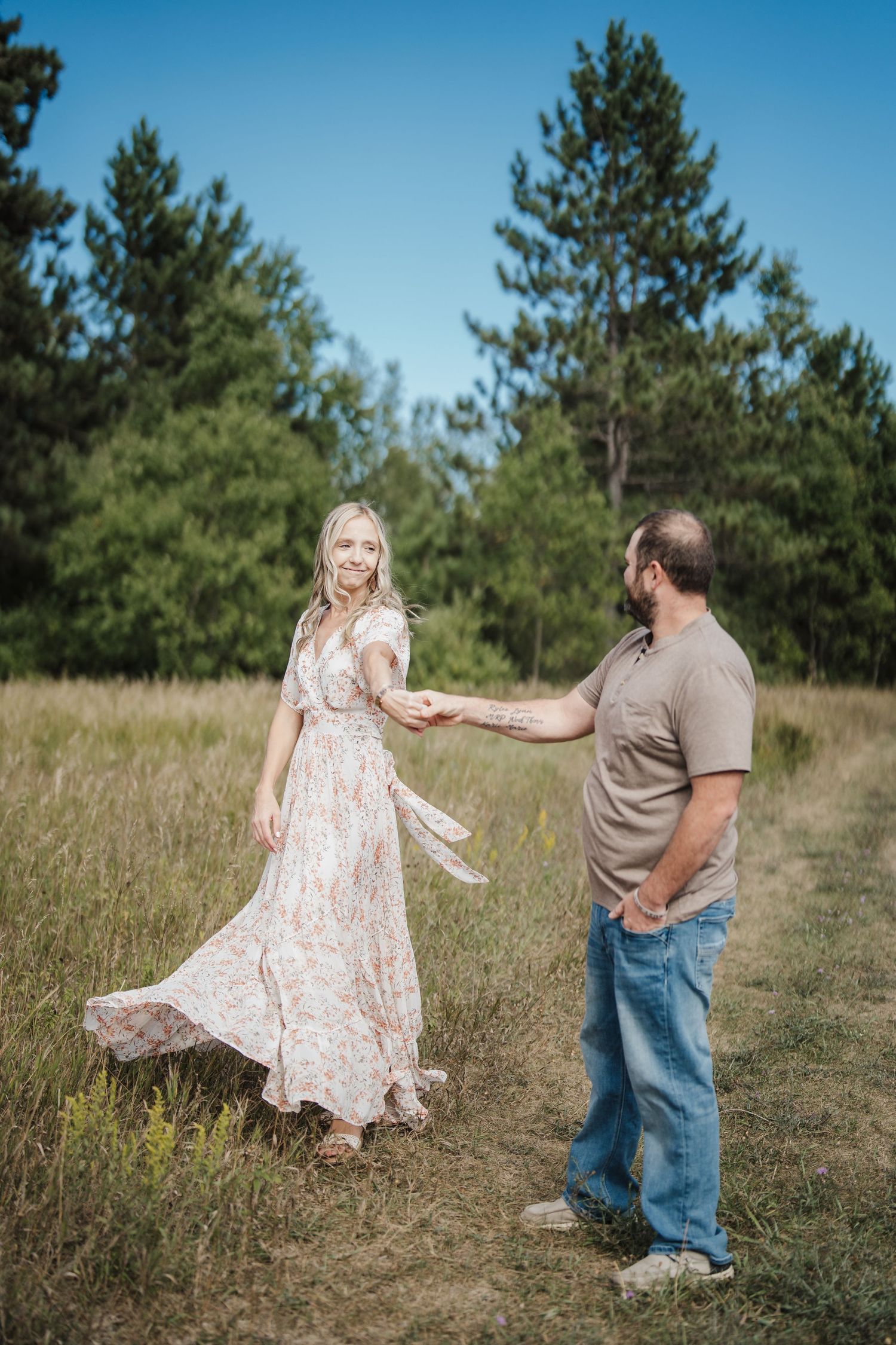 A couple dances playfully together in a sunlit meadow with pine trees in the background.
