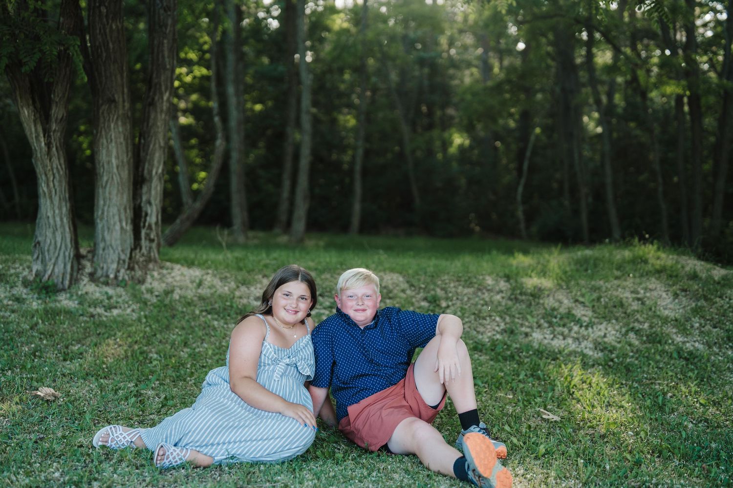 A couple sits together on green grass in a serene wooded setting during a summer afternoon.