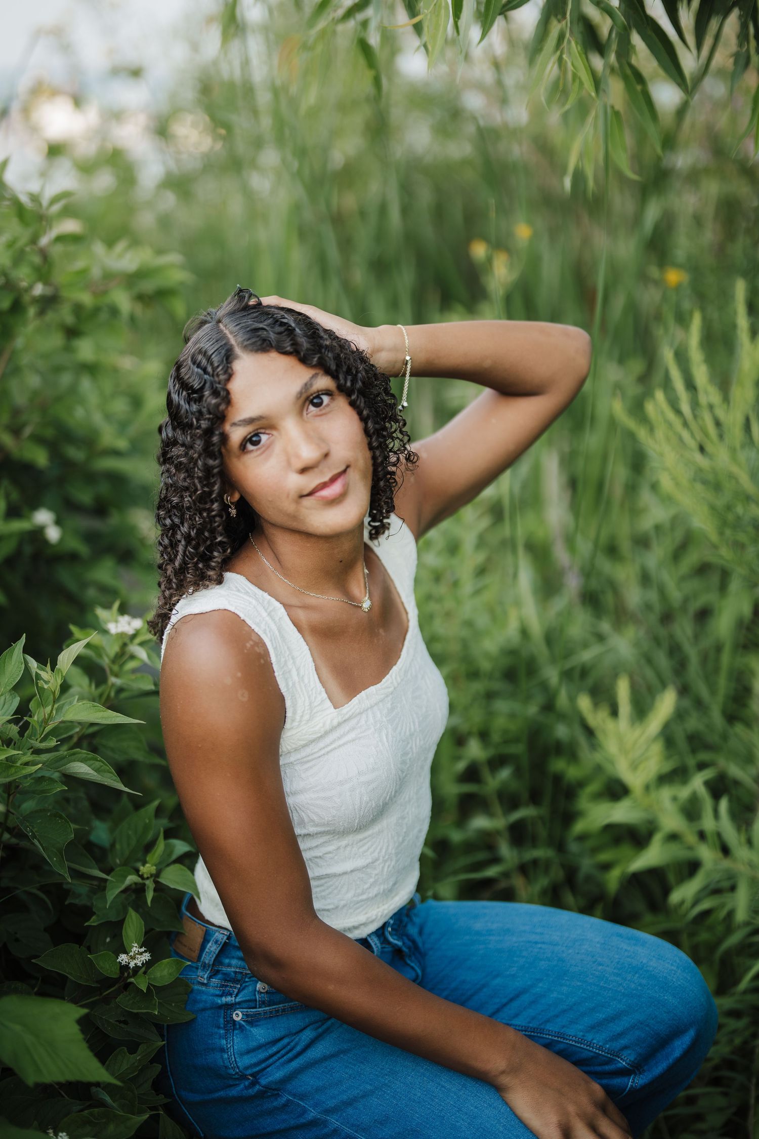 Outdoor portrait session in lush greenery featuring casual summer style with white tank top and blue jeans.