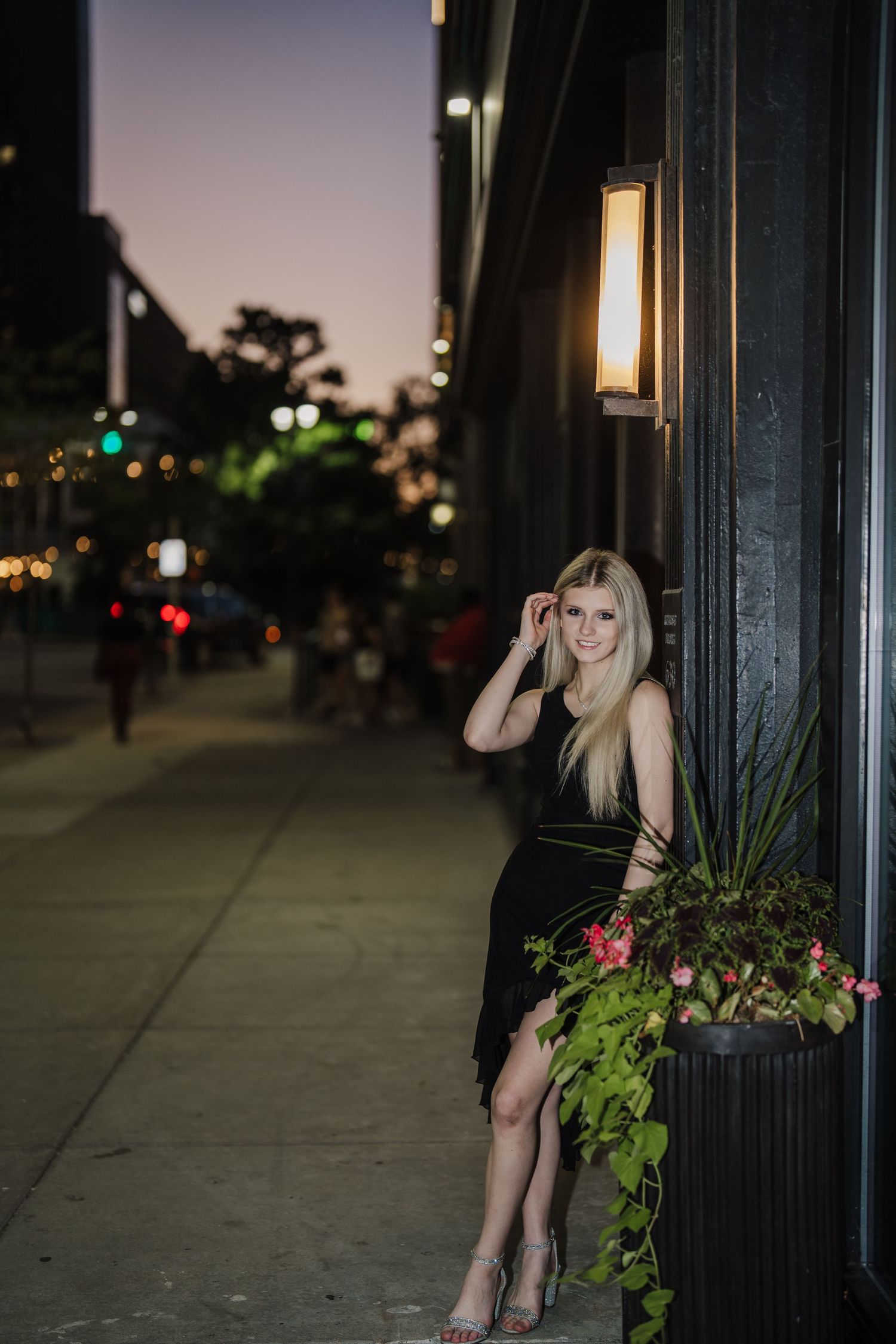 Woman in black dress poses elegantly near illuminated street lamp and decorative planter at night.