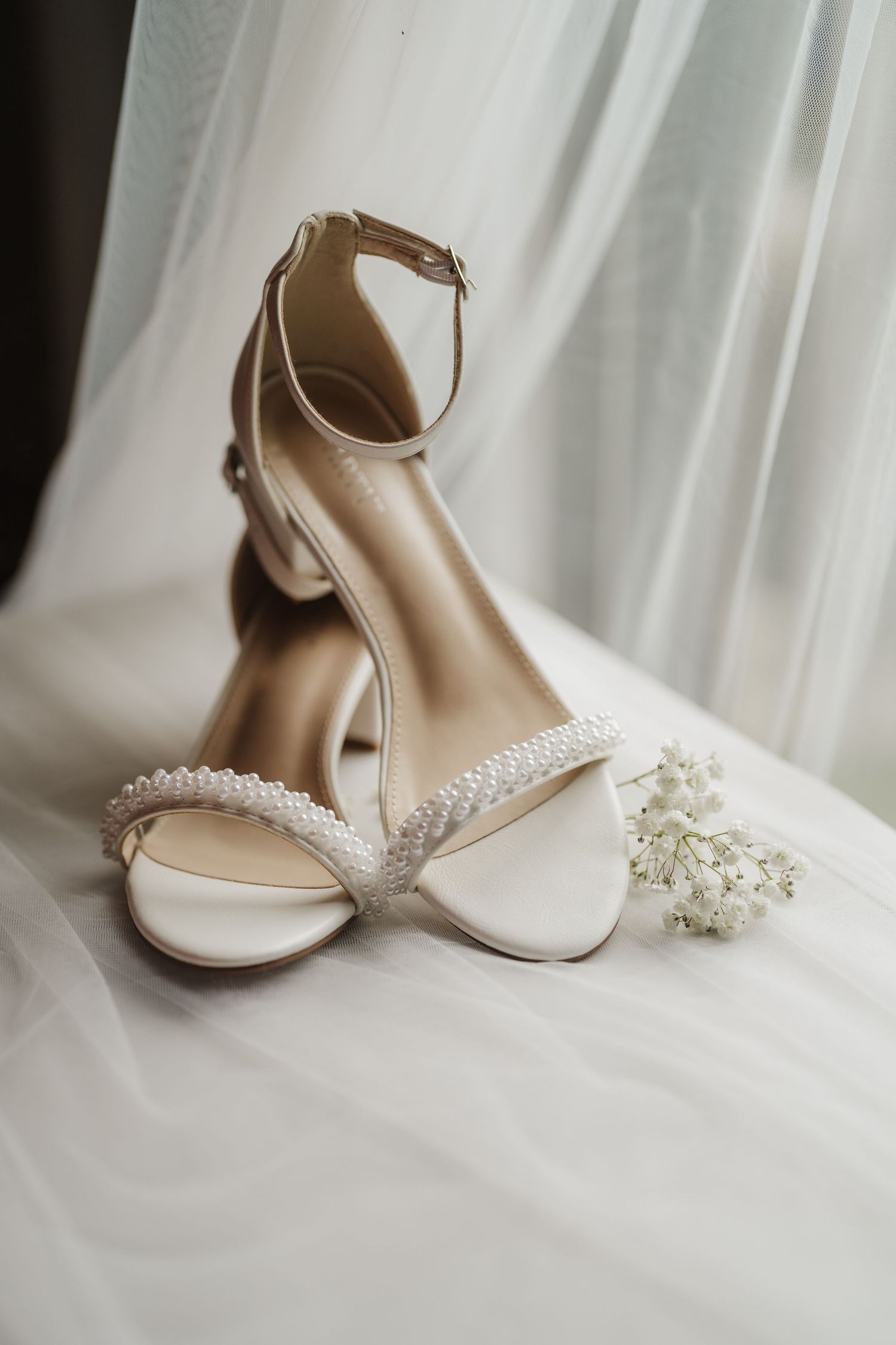Elegant white bridal heels with pearl embellishments and ankle straps displayed against a soft white backdrop.