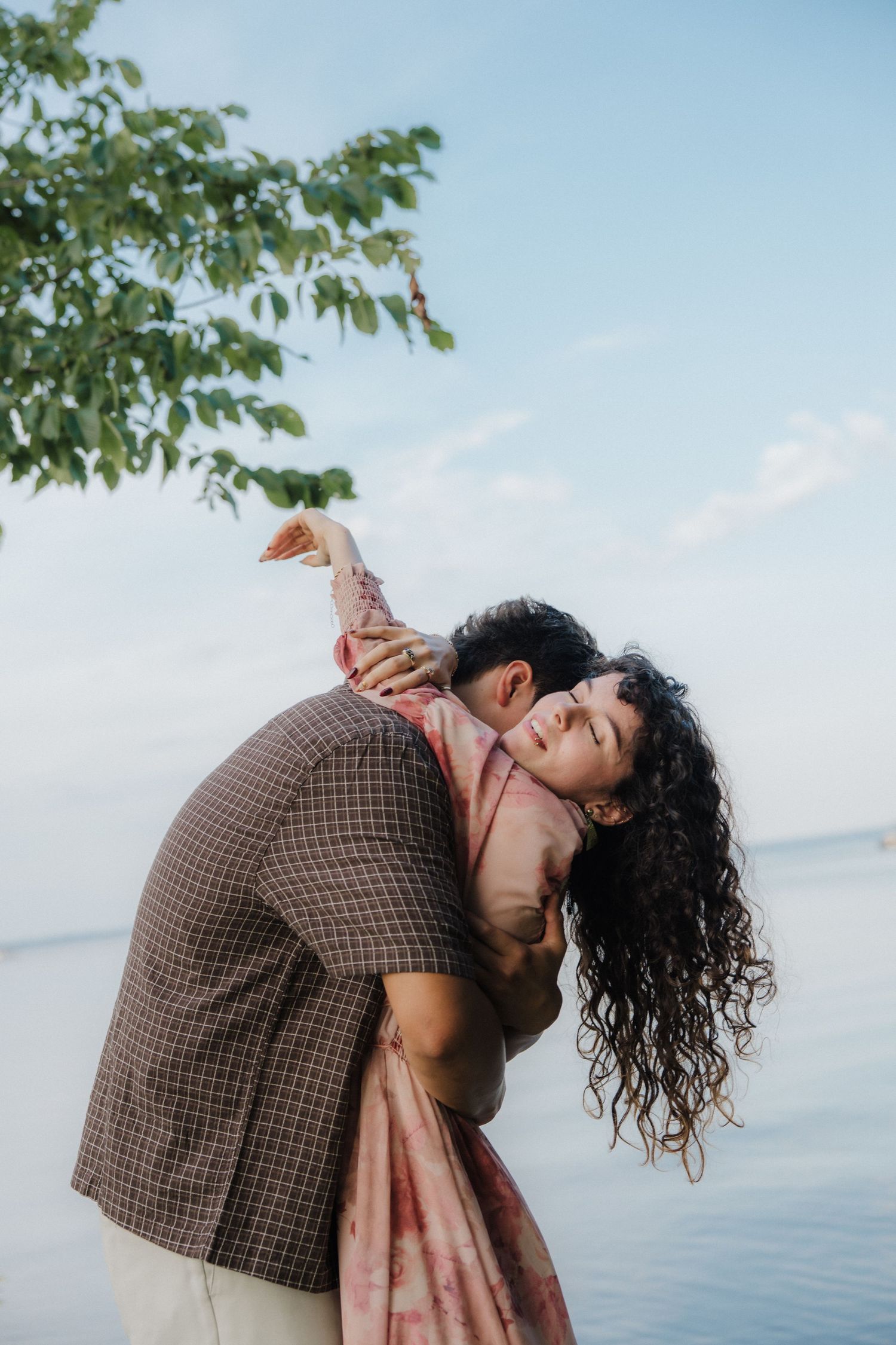 Romantic couple dancing by the water, man dipping woman in floral dress near a tree with blue sky backdrop.