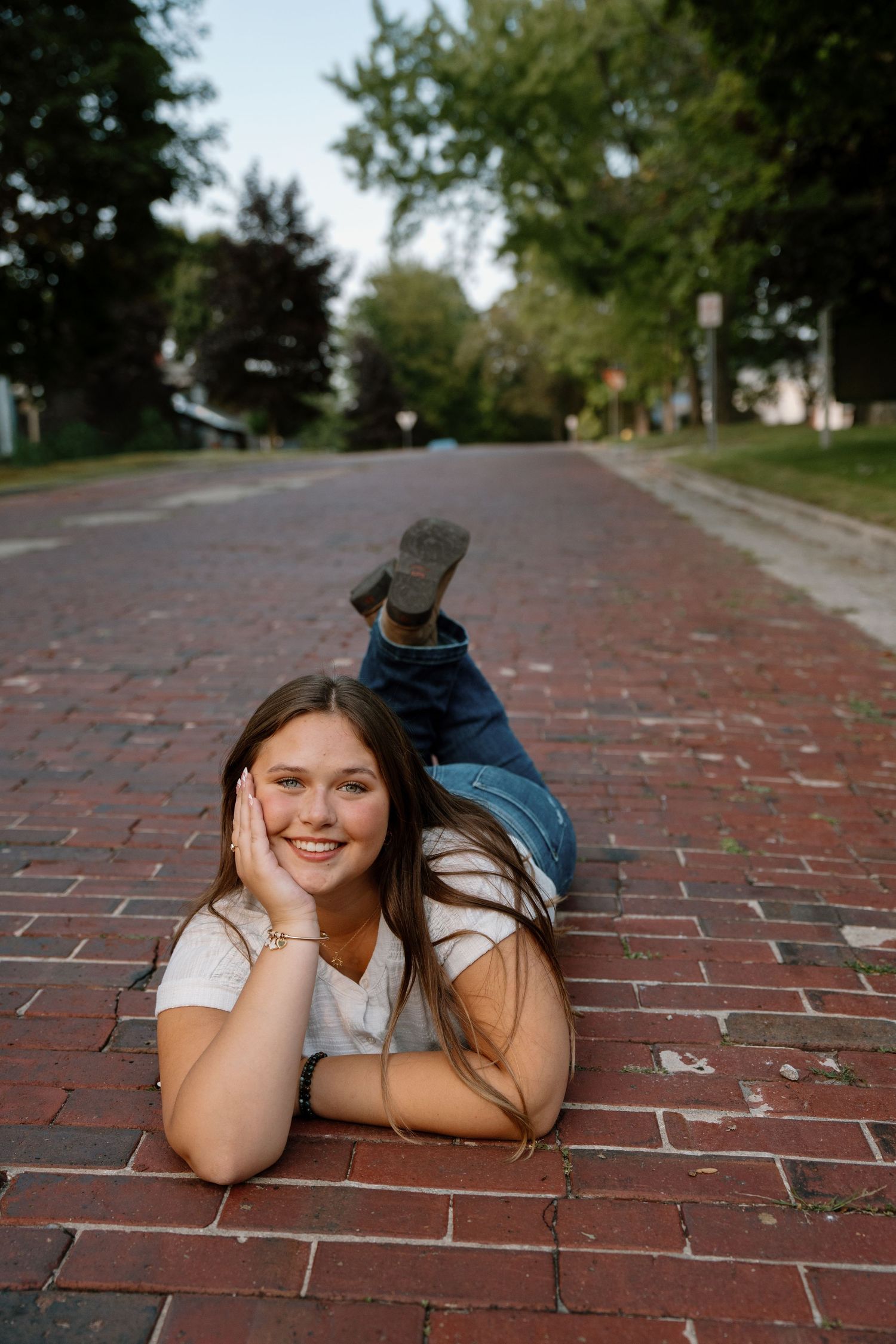 Young woman in jeans and white top lying on a brick road, smiling with hand on cheek, trees in background.
