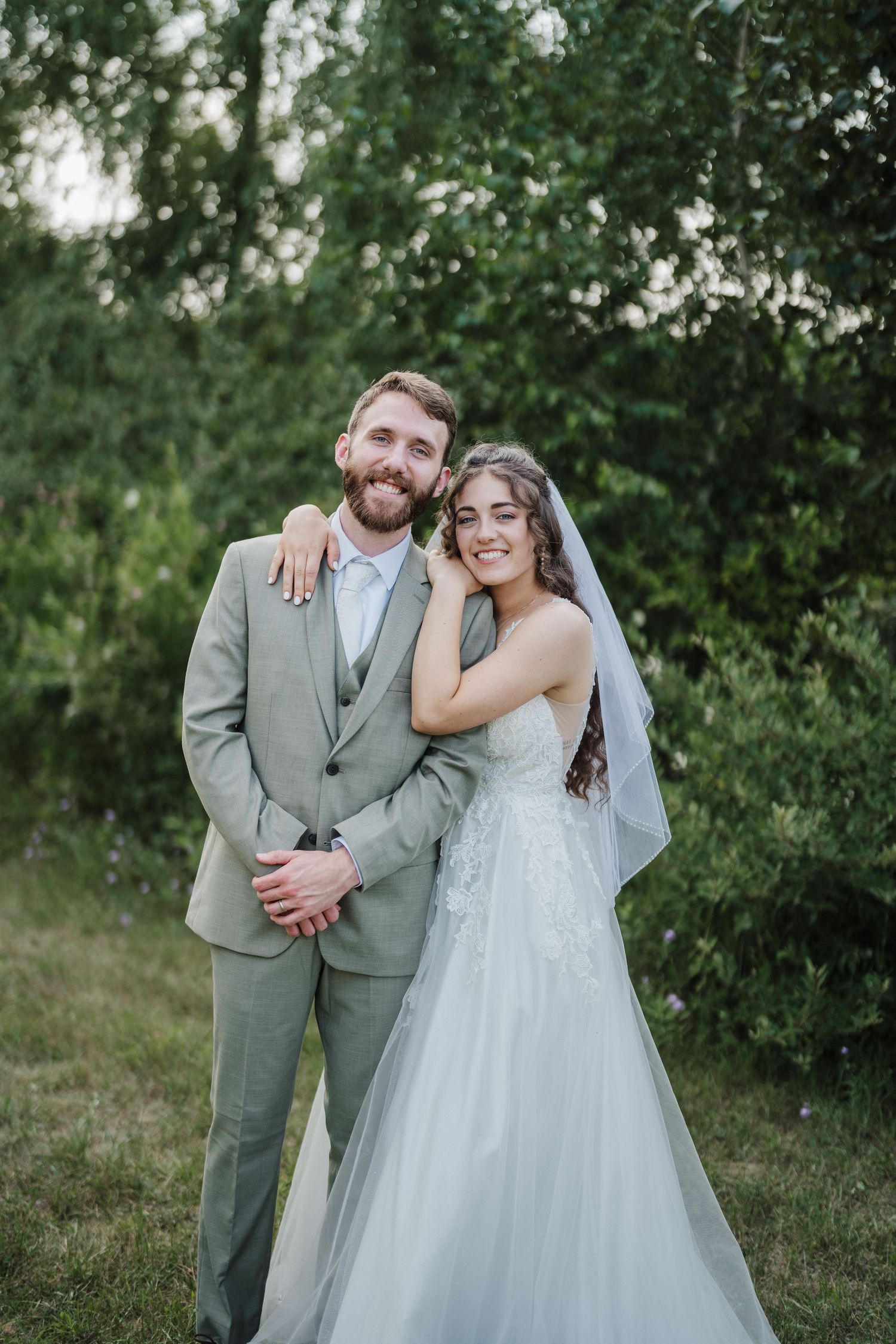 Newlyweds pose together outdoors, bride in white lace gown and groom in light grey suit.
