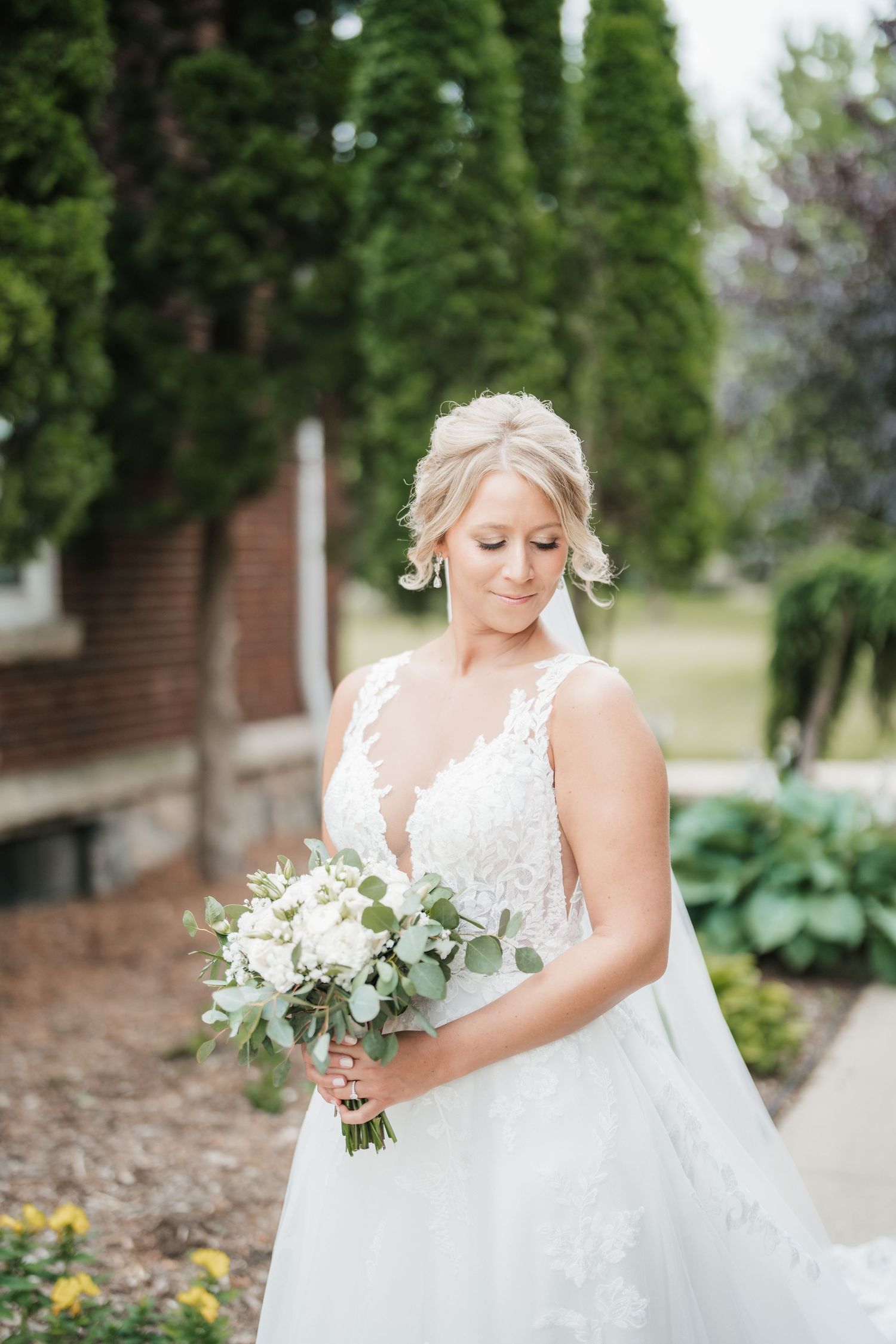 Bride in a white lace gown holds a beautiful bouquet of white flowers in an outdoor garden setting.