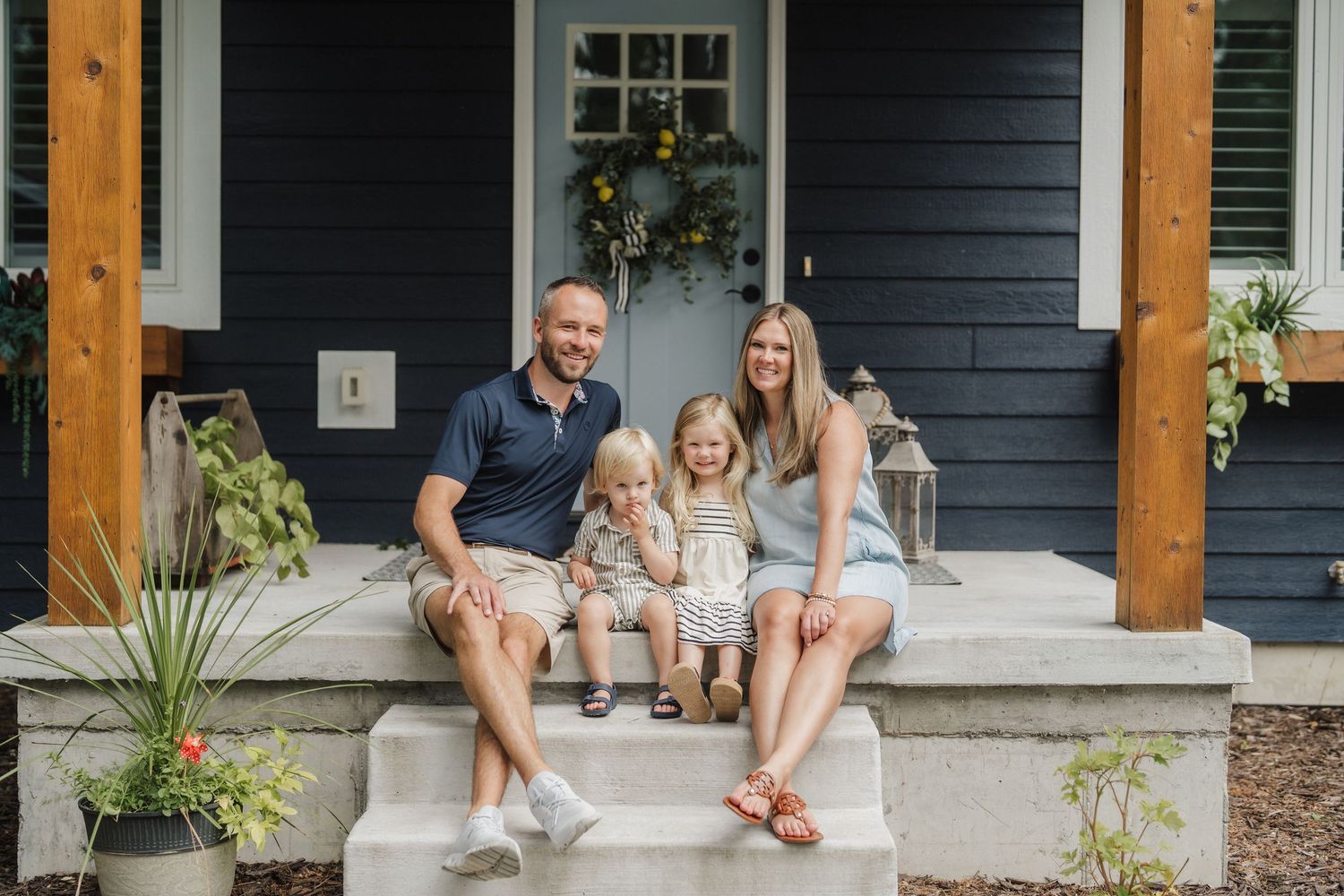 Happy family of four sitting on front porch steps of a navy blue craftsman home on a sunny day.