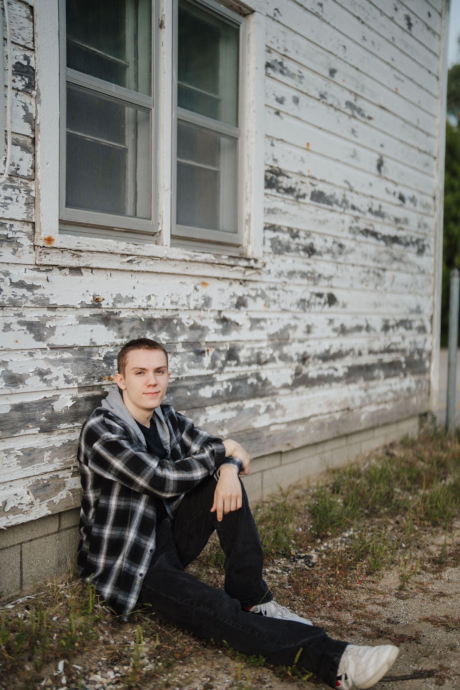 A person in a plaid flannel shirt and dark pants sits against a weathered white building exterior.