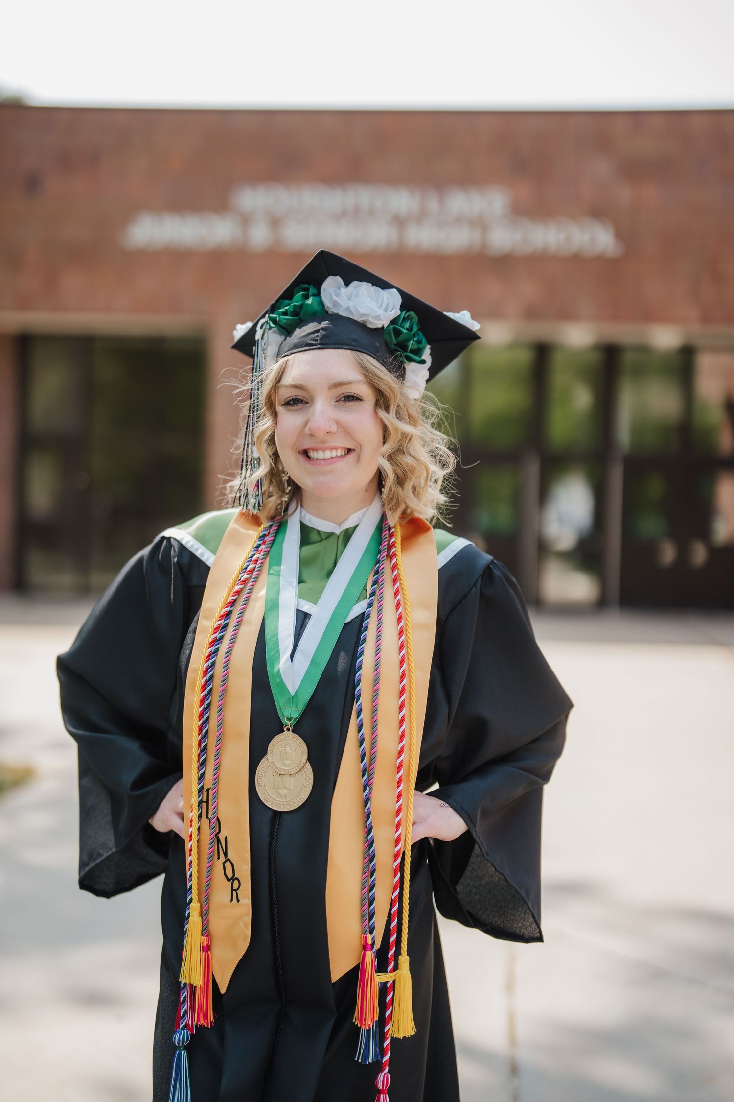 Graduate in black cap and gown with colorful cords and medallion poses outside academic building.
